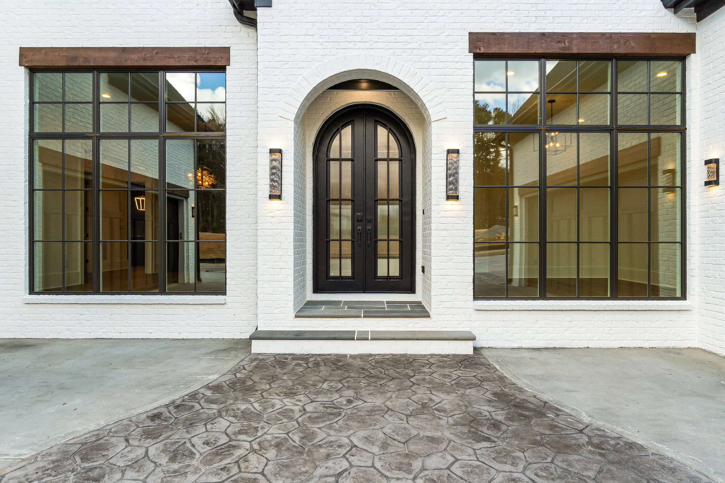 Front view of a modern house with white brick exterior, black front door with glass panels, large windows on either side, and brick and stone walkways leading up to the entrance.