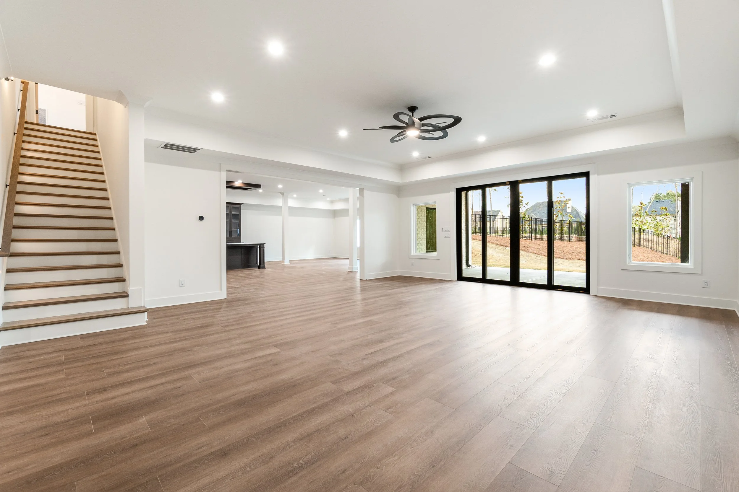 Empty living room with hardwood floors, white walls, large sliding glass doors leading outside, and a staircase with wooden steps on the left. Ceiling fan and recessed lighting are installed.