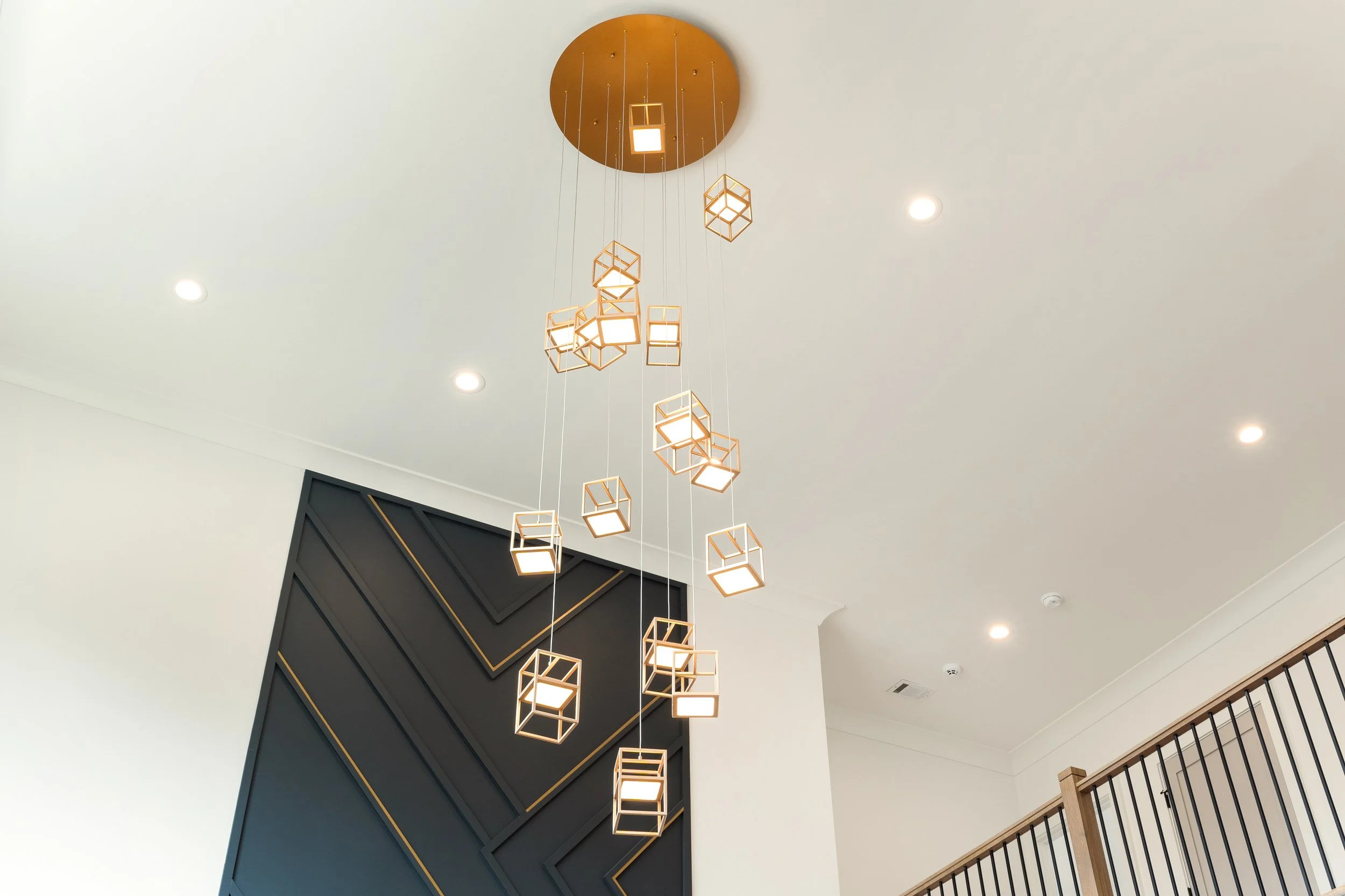 Modern hanging chandelier with geometric cube-shaped frames and glowing lights, mounted on a circular ceiling fixture in a high-ceiling room with recessed lighting and a black accent wall.