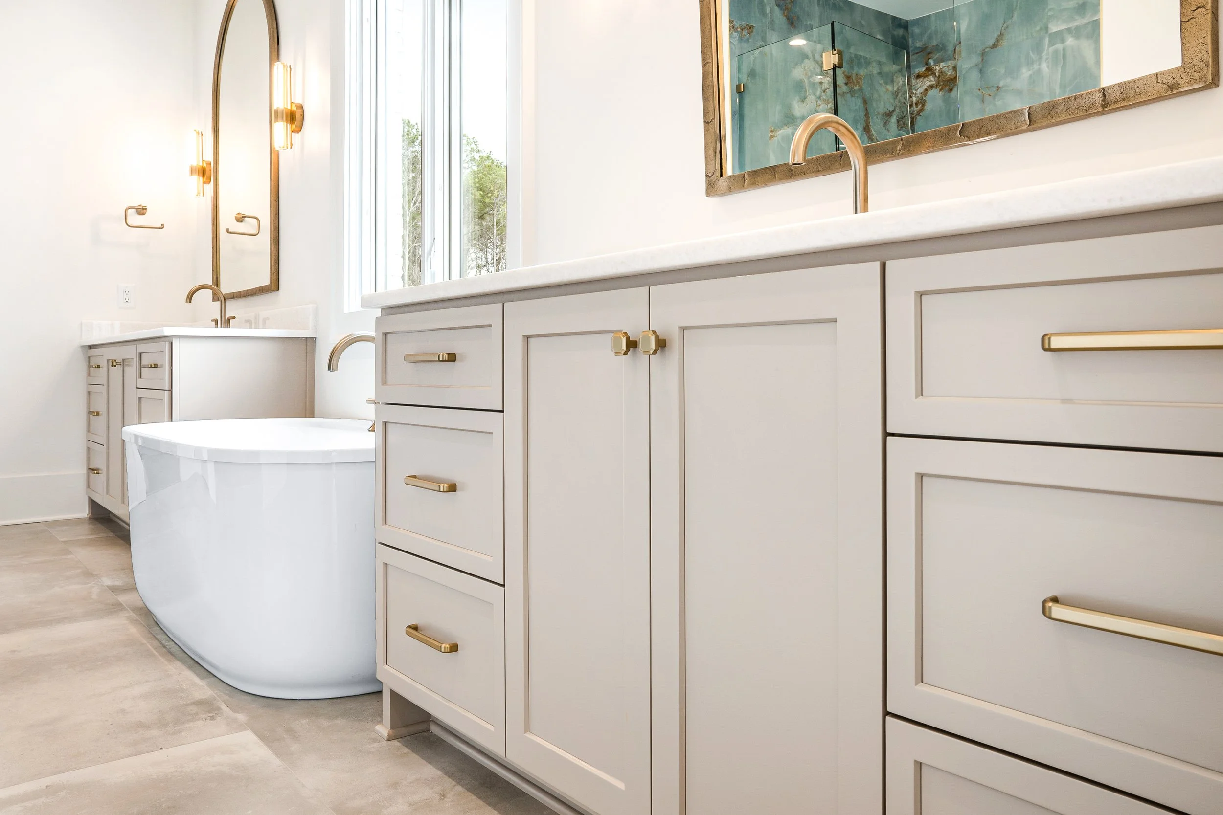 A modern bathroom with white cabinetry, gold hardware, a marble countertop, and a large mirror above the sink. There is a glass-enclosed shower with blue marble tiles and a bathtub beside the cabinetry. Natural light enters through a tall window.