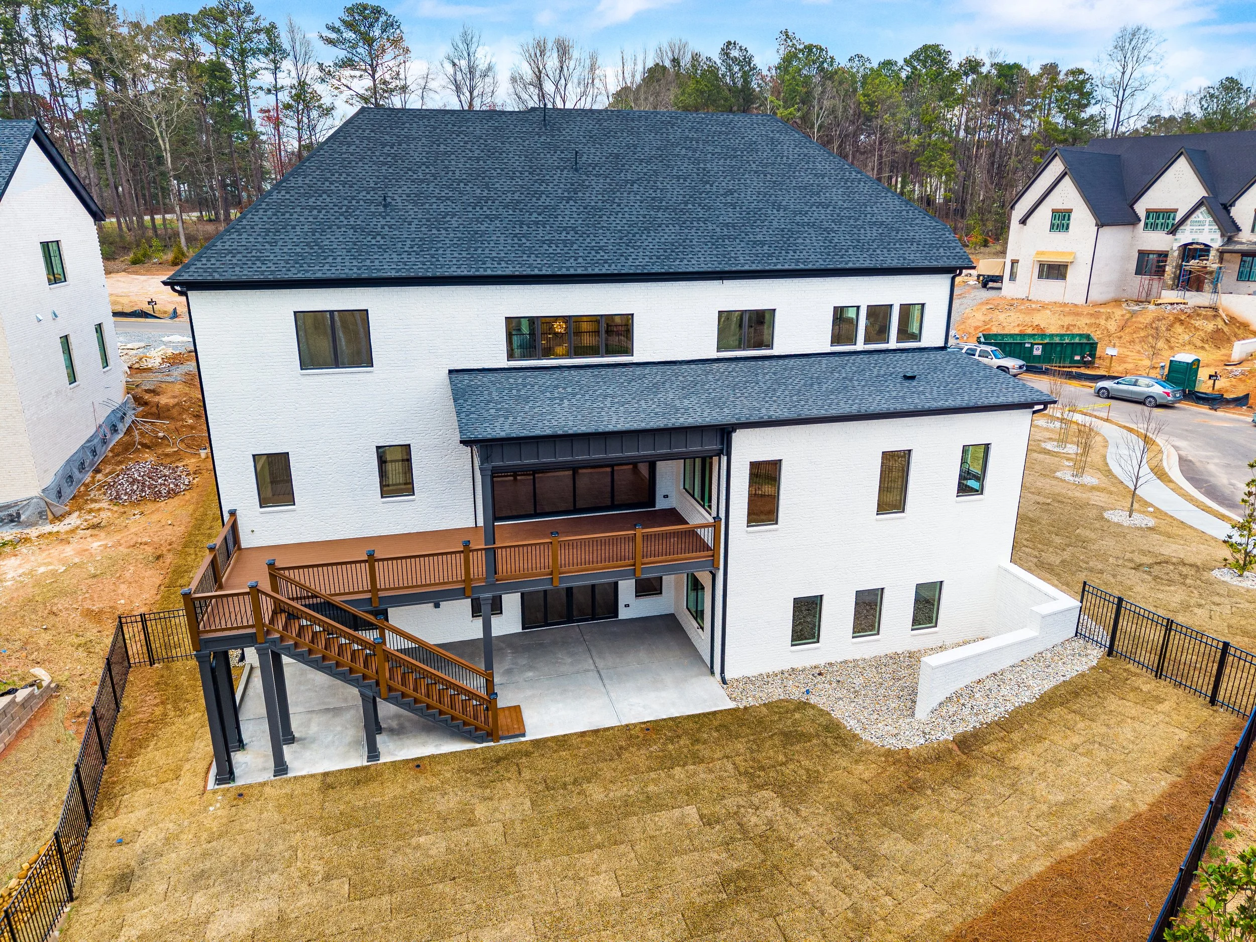 Aerial view of a new white house with black roofing, large windows, a wooden balcony, and fenced yard under an overcast sky, with neighboring houses and trees nearby.