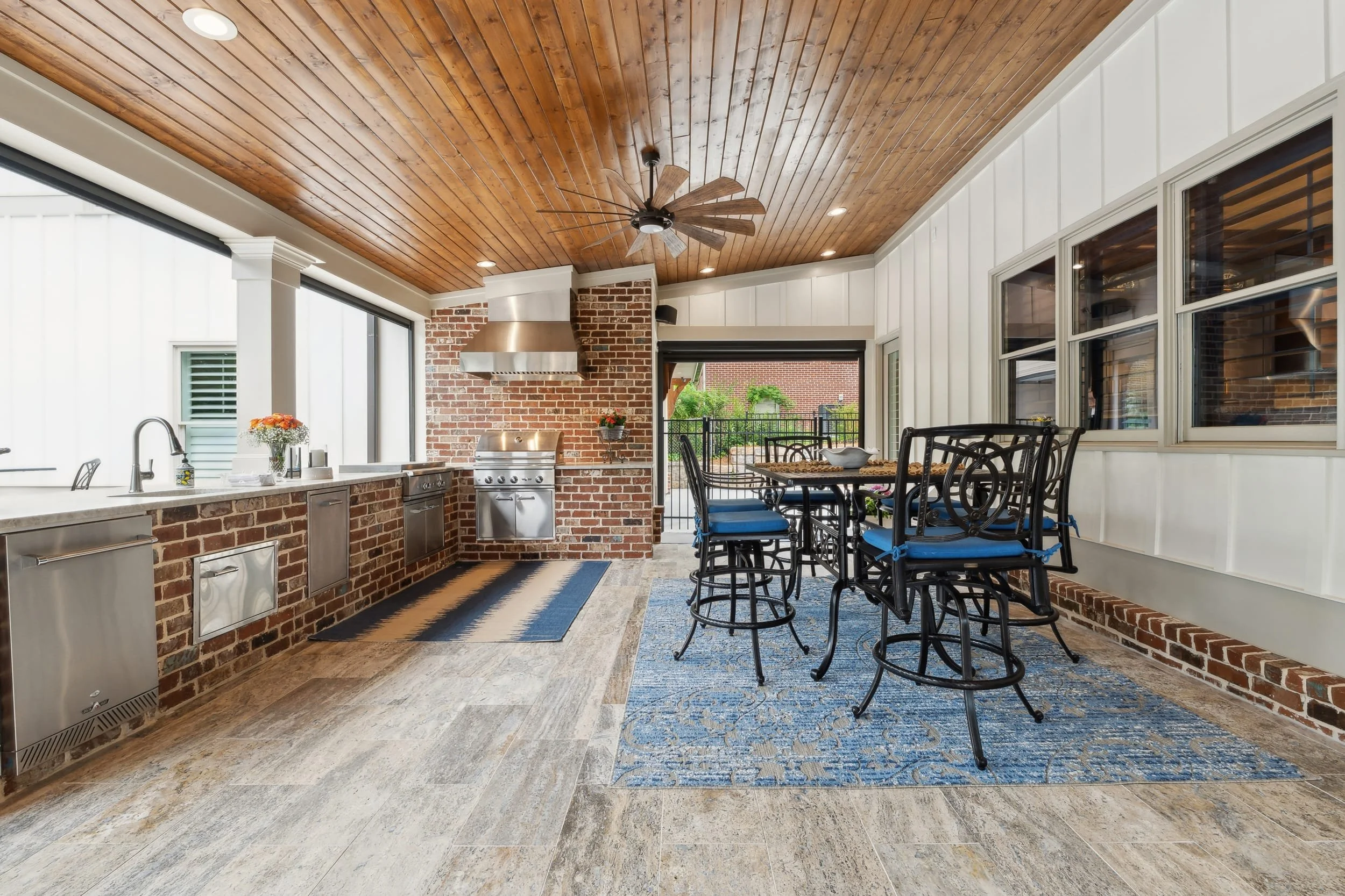 Indoor patio with a wooden ceiling, brick walls, a stainless steel grill, black metal dining table with six chairs, large windows, and a blue patterned rug.