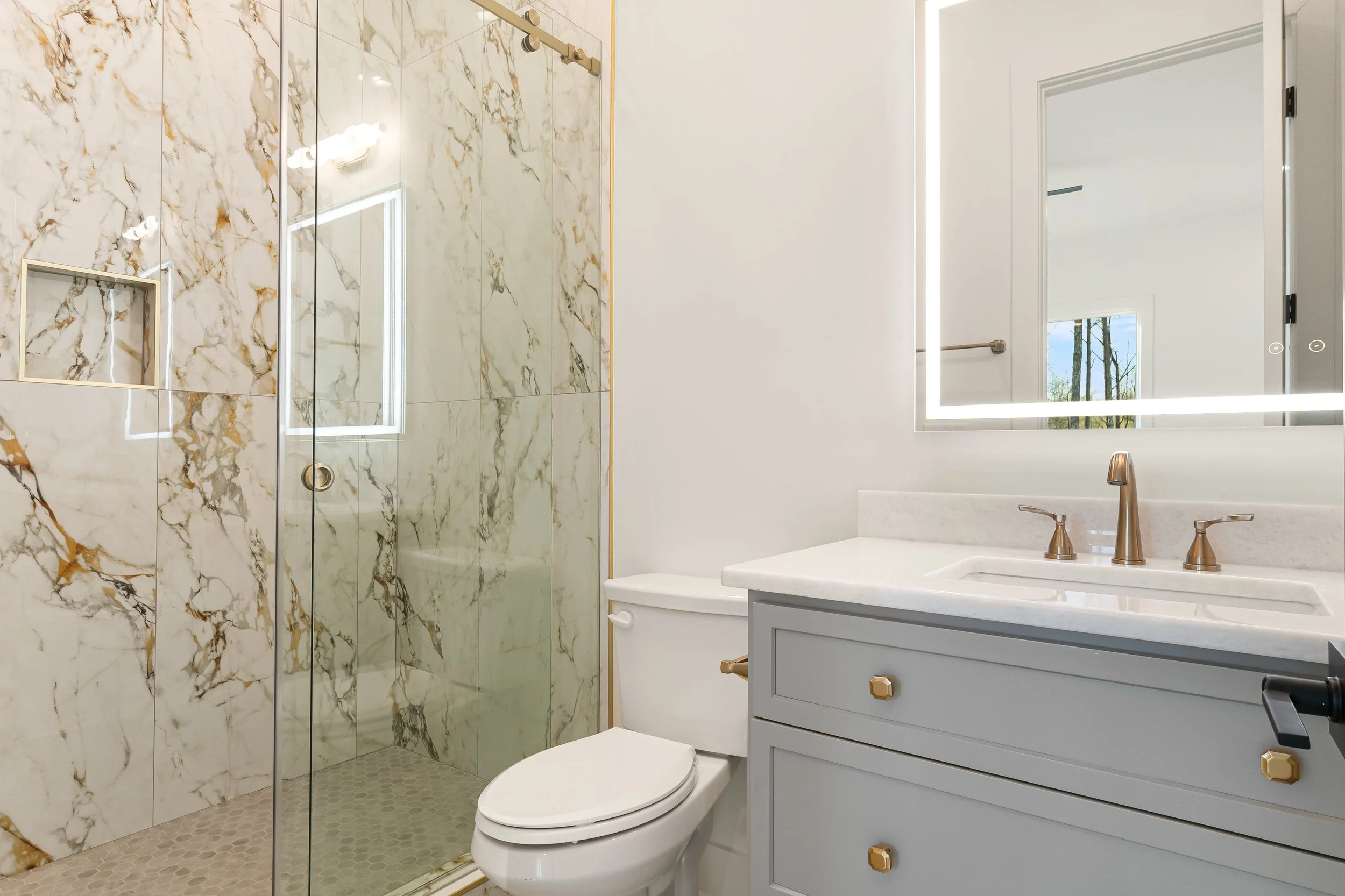 A modern bathroom featuring a marble-tiled shower with a built-in niche, a large mirror with LED lighting, a light blue vanity with gold knobs, a white countertop with a gold faucet, and a window showing trees outside.