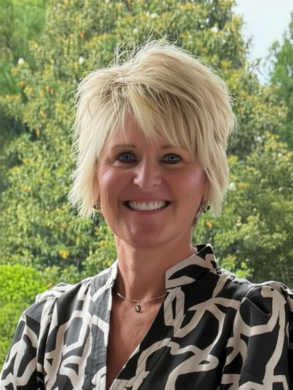 A woman with short blonde hair smiling outdoors with trees in the background.
