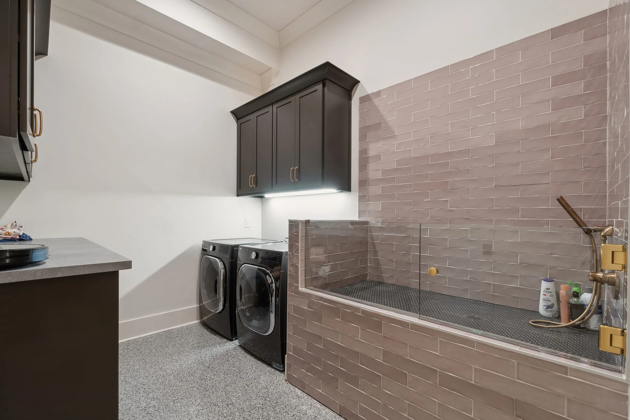 A laundry room with dark cabinetry, a washing machine and dryer, a tiled section with a built-in tub, and various toiletries and cleaning supplies.