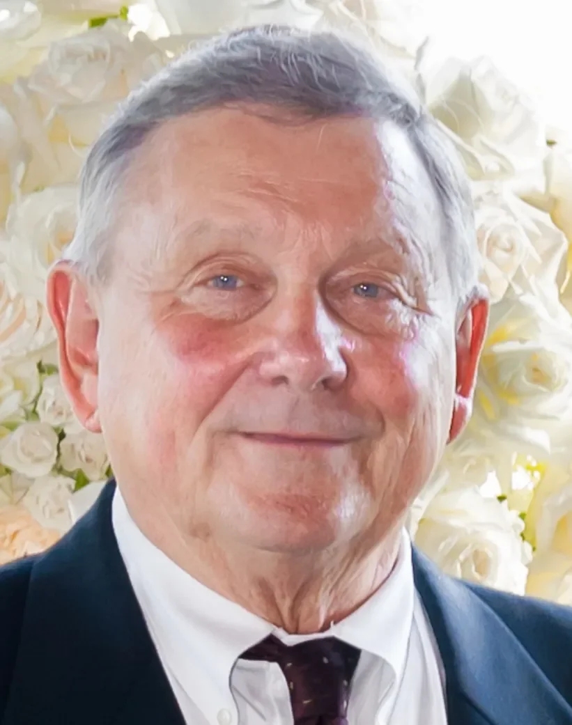 Older man with gray hair and blue eyes wearing a black tuxedo, white dress shirt, and black tie, standing in front of a background filled with white roses.
