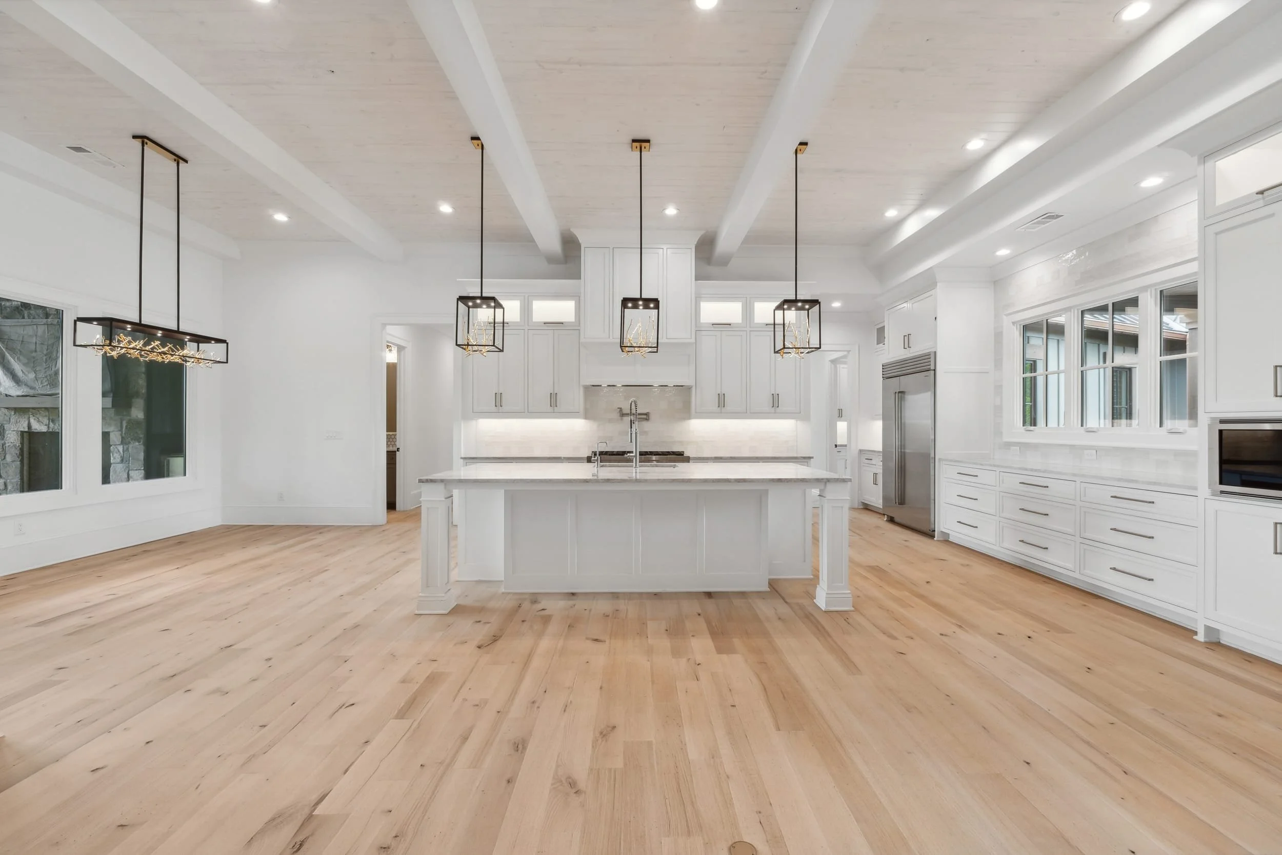 Bright modern kitchen with white cabinetry, marble island, and pendant lighting fixtures, featuring hardwood floors and large windows.
