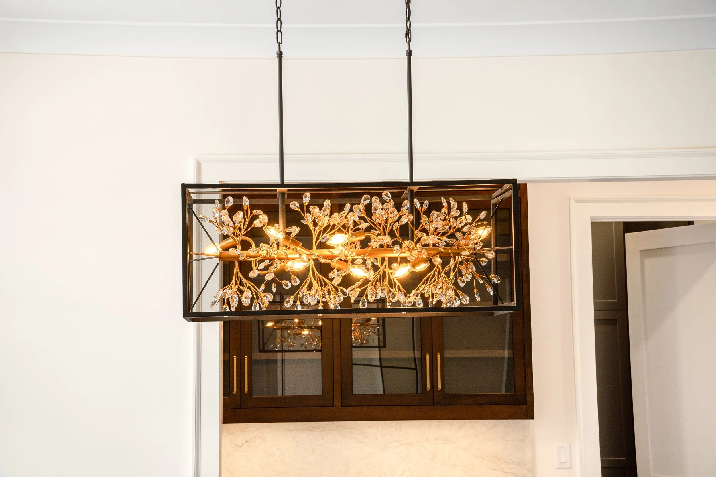 A modern chandelier with crystal accents hanging from black metal frame in a room with white walls and a mirror reflection.