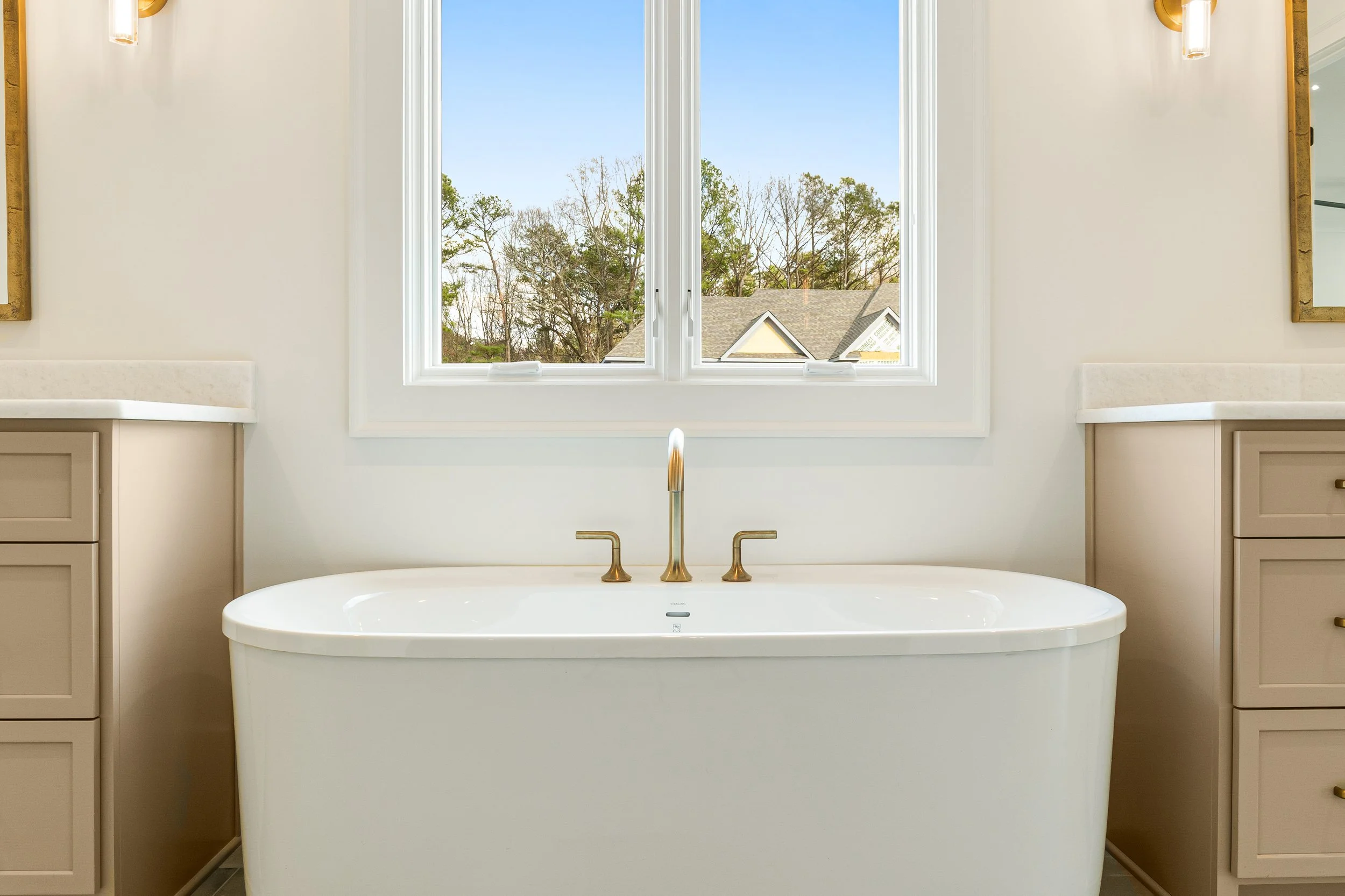 A white bathtub with gold fixtures positioned below a large window with a view of trees and rooftops in a bright, modern bathroom.