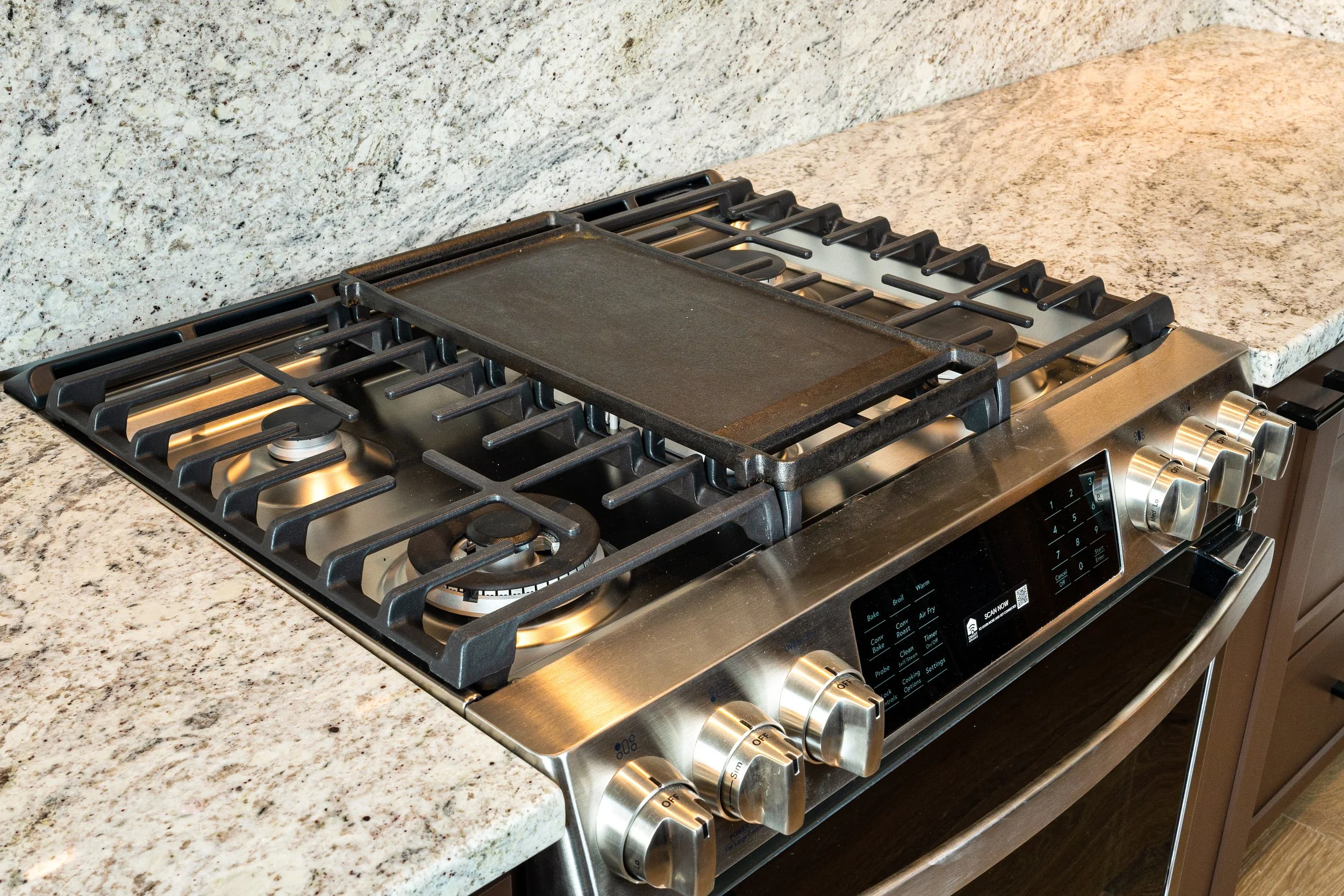 A stainless steel gas stove with four burners and a black cast iron griddle on top, set into a granite countertop.