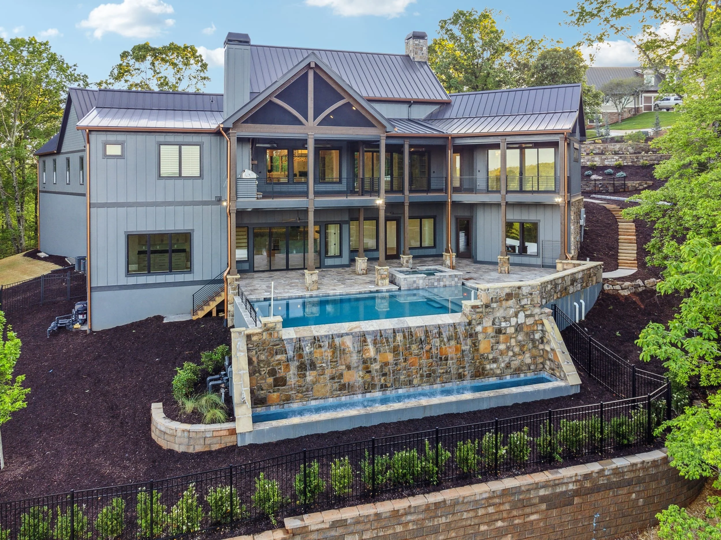 A modern, multi-story house with a metal roof, large windows, a spacious balcony, and a backyard swimming pool. The house is surrounded by trees and landscaped garden, with a hillside and additional houses in the background.