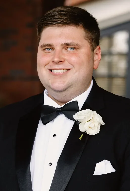 Portrait of a man in a black tuxedo with a white shirt and black bow tie, smiling, with a white boutonniere on his left lapel, indoors with blurred background.