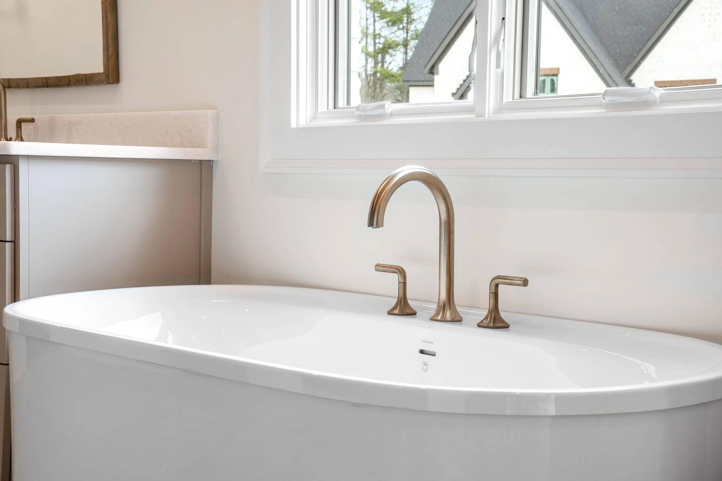 A white freestanding bathtub with a shiny bronze faucet and handles, situated near a window with a view of neighboring houses.