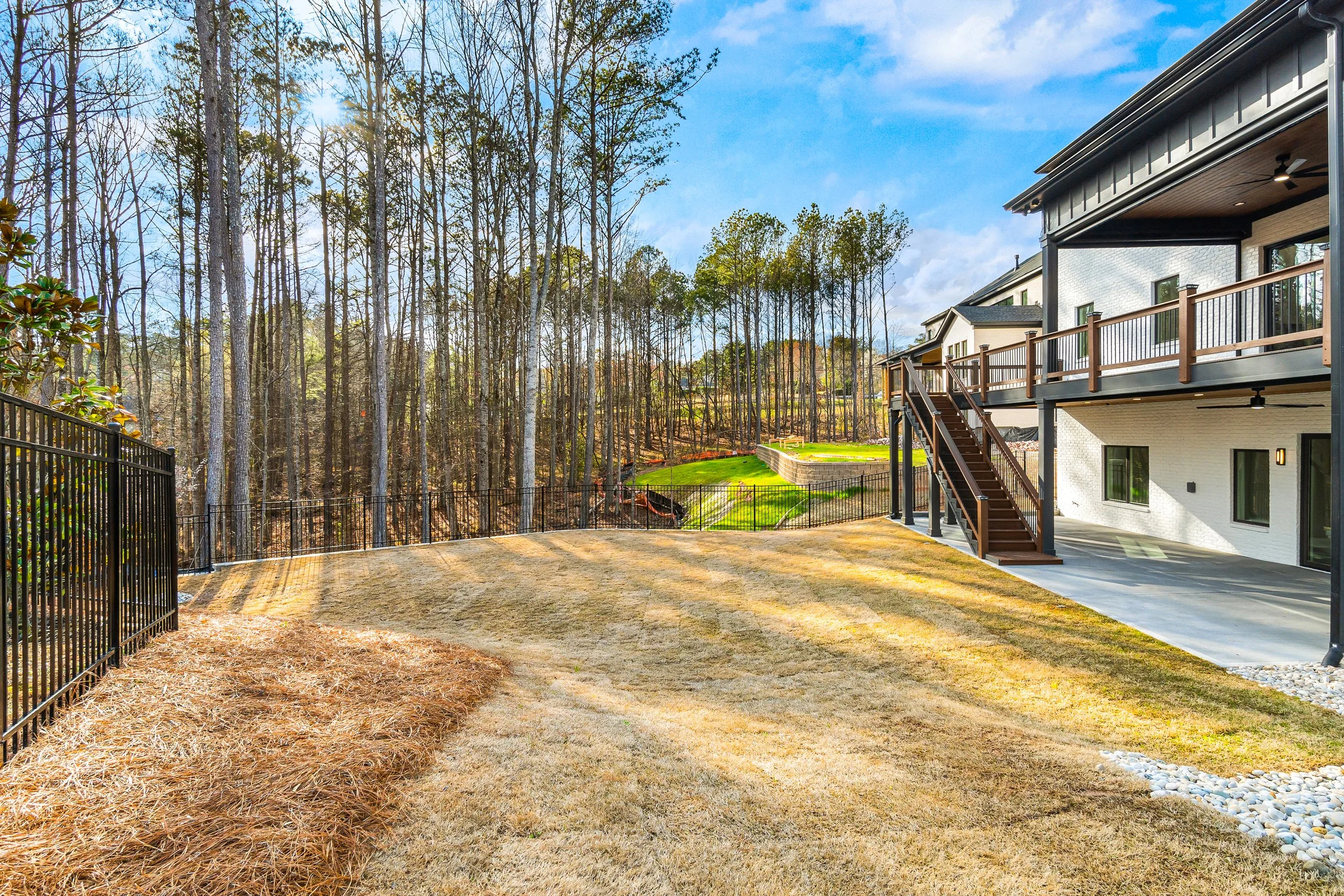 Backyard view with new house, wooden deck, stairs, fenced yard, grassy area, and trees in the background under a blue sky with some clouds.