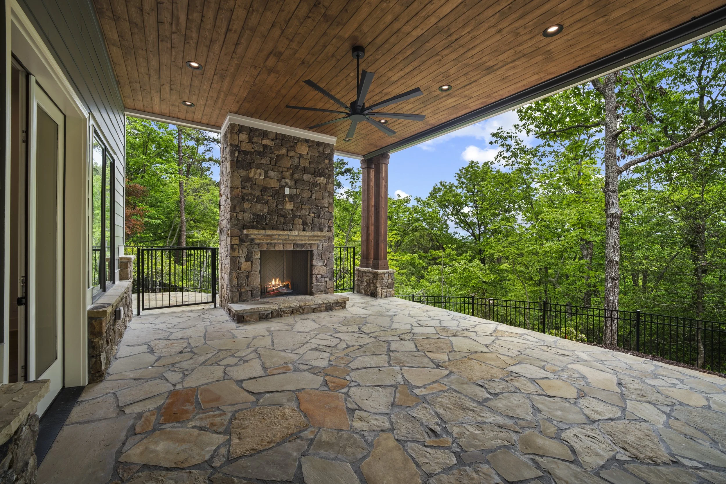 Covered outdoor patio with stone flooring, a stone fireplace with a fire burning, a ceiling fan, recessed lighting, and surrounded by lush green trees.
