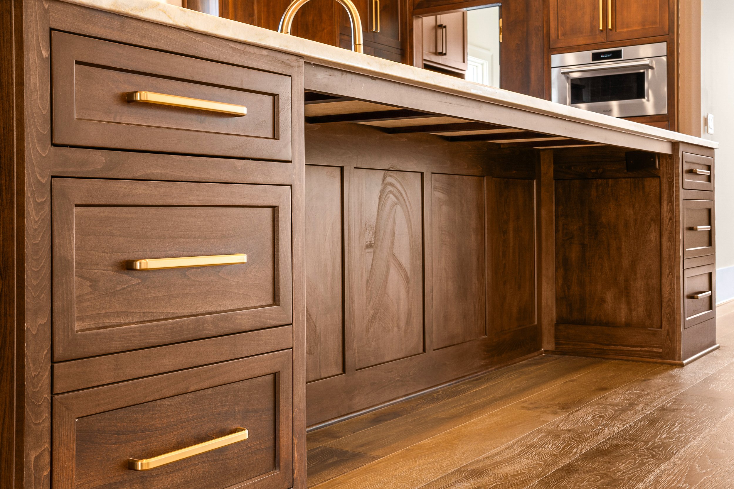 A kitchen with dark wooden cabinets, a cream-colored stone countertop, and a built-in stainless steel microwave oven. The floor is made of matching dark wood.