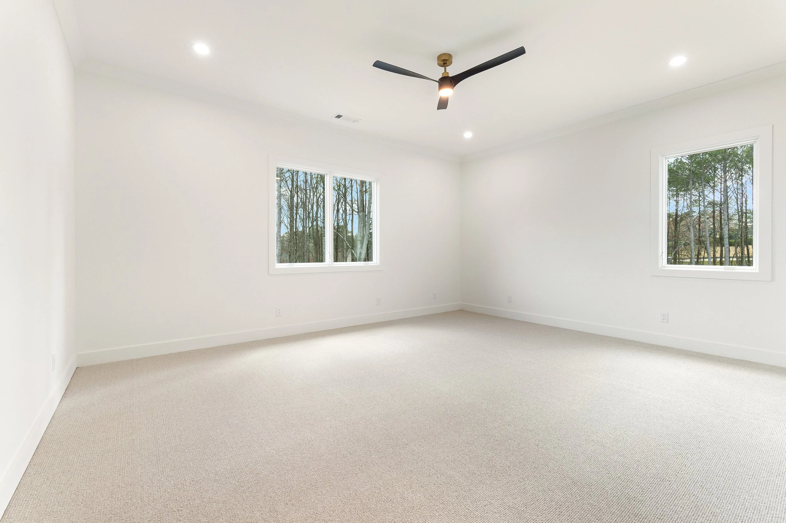 Empty room with white walls, beige carpet, two windows showing trees outside, and a black ceiling fan with three blades and a light in the center.