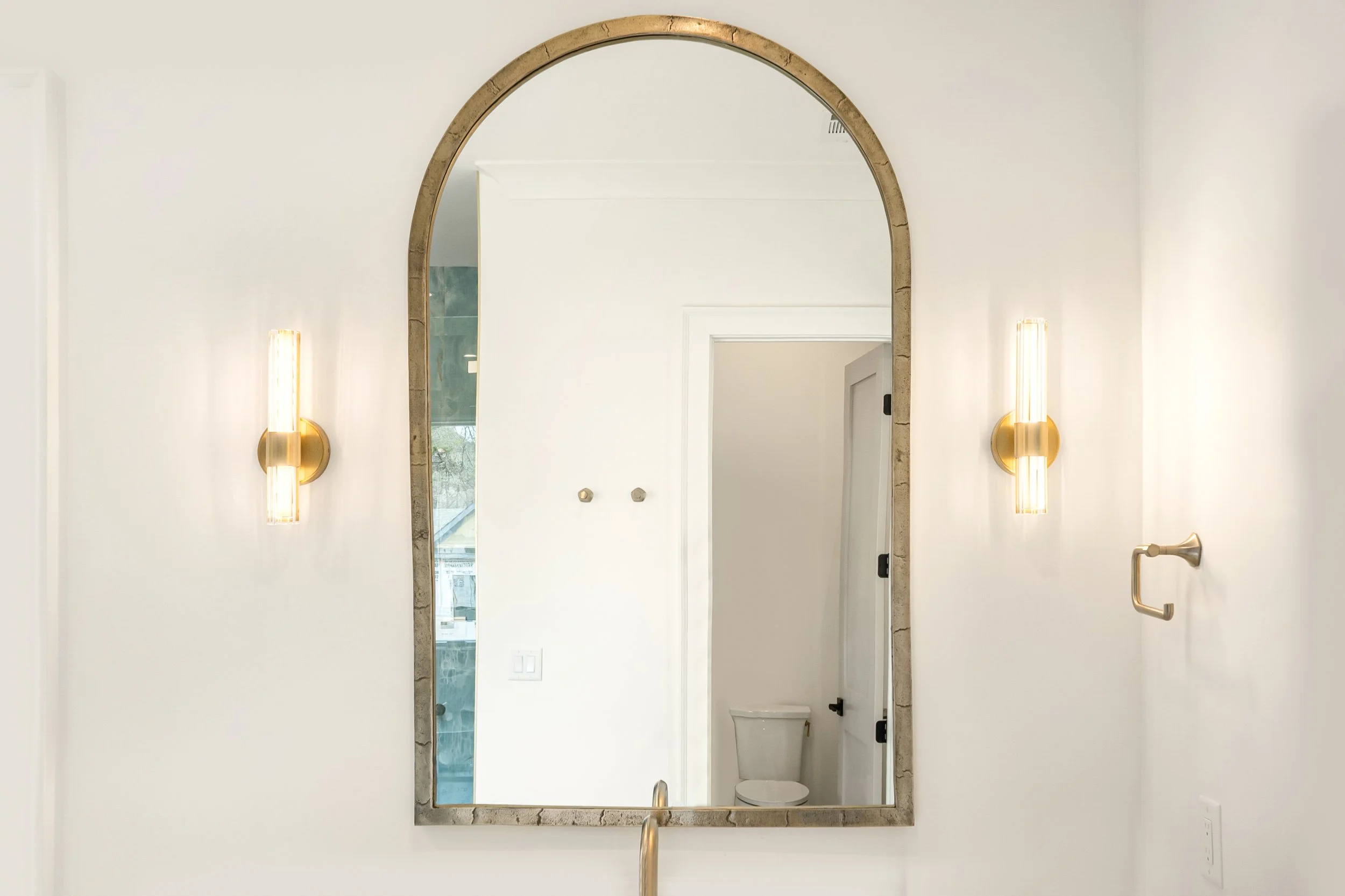 A large mirror with an arched top, framed in wood, mounted on a white wall. Two wall-mounted brass and glass light fixtures flank the mirror. The reflection shows part of a bathroom with a white door, a toilet, and a window.