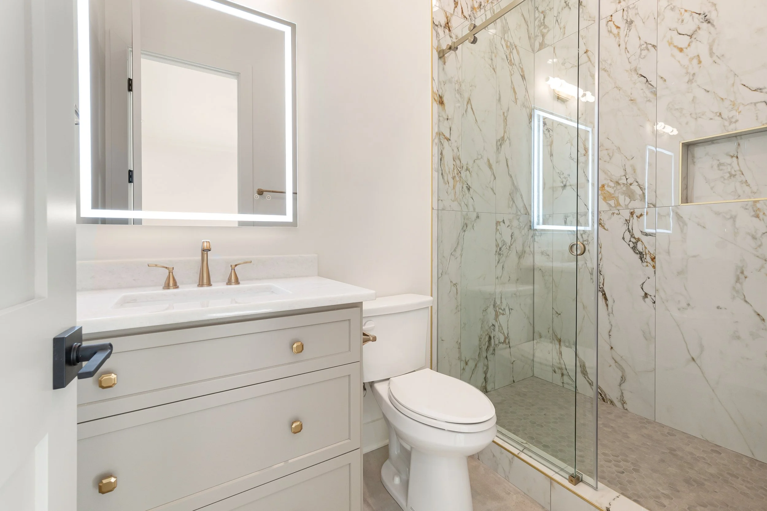 A modern bathroom featuring a white vanity with gold hardware, a large illuminated mirror, a marble shower with glass door, and marble walls with gold veining.