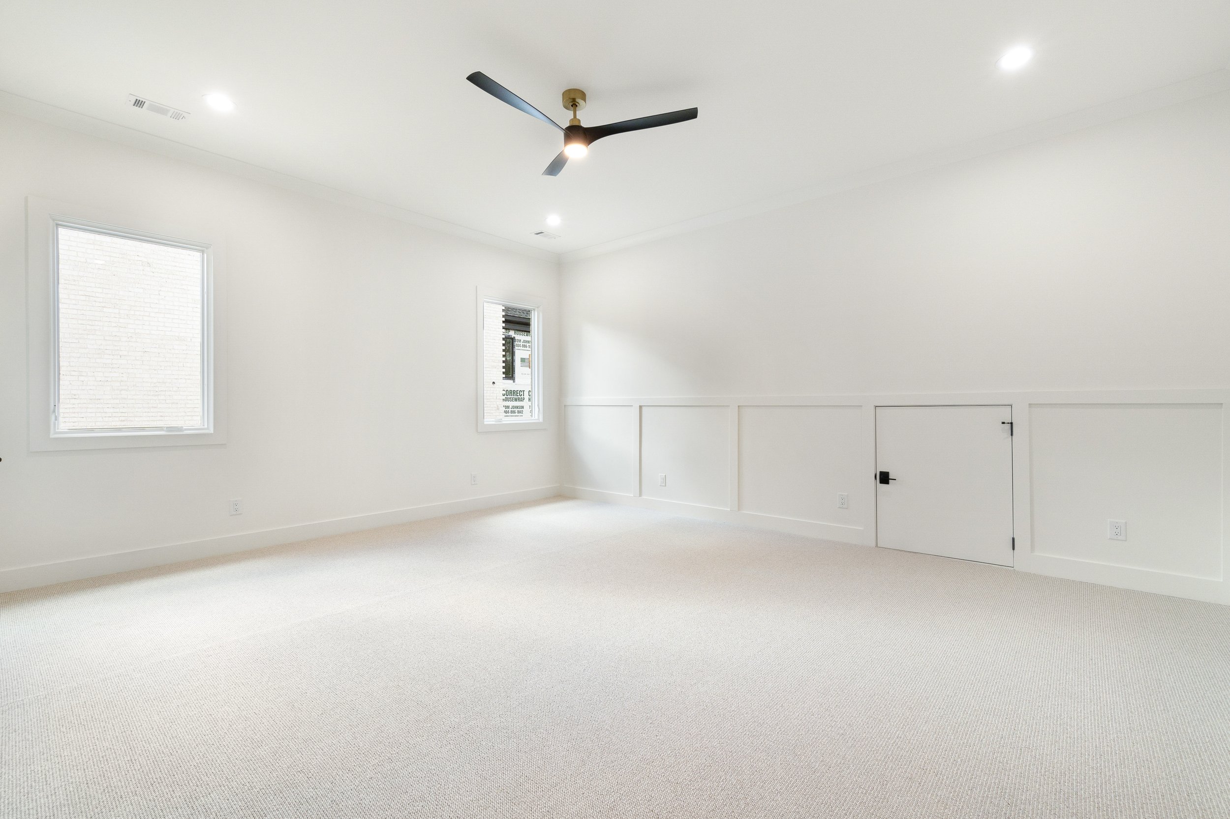 Empty room with white walls, beige carpet, two windows, a small door, and a ceiling fan.