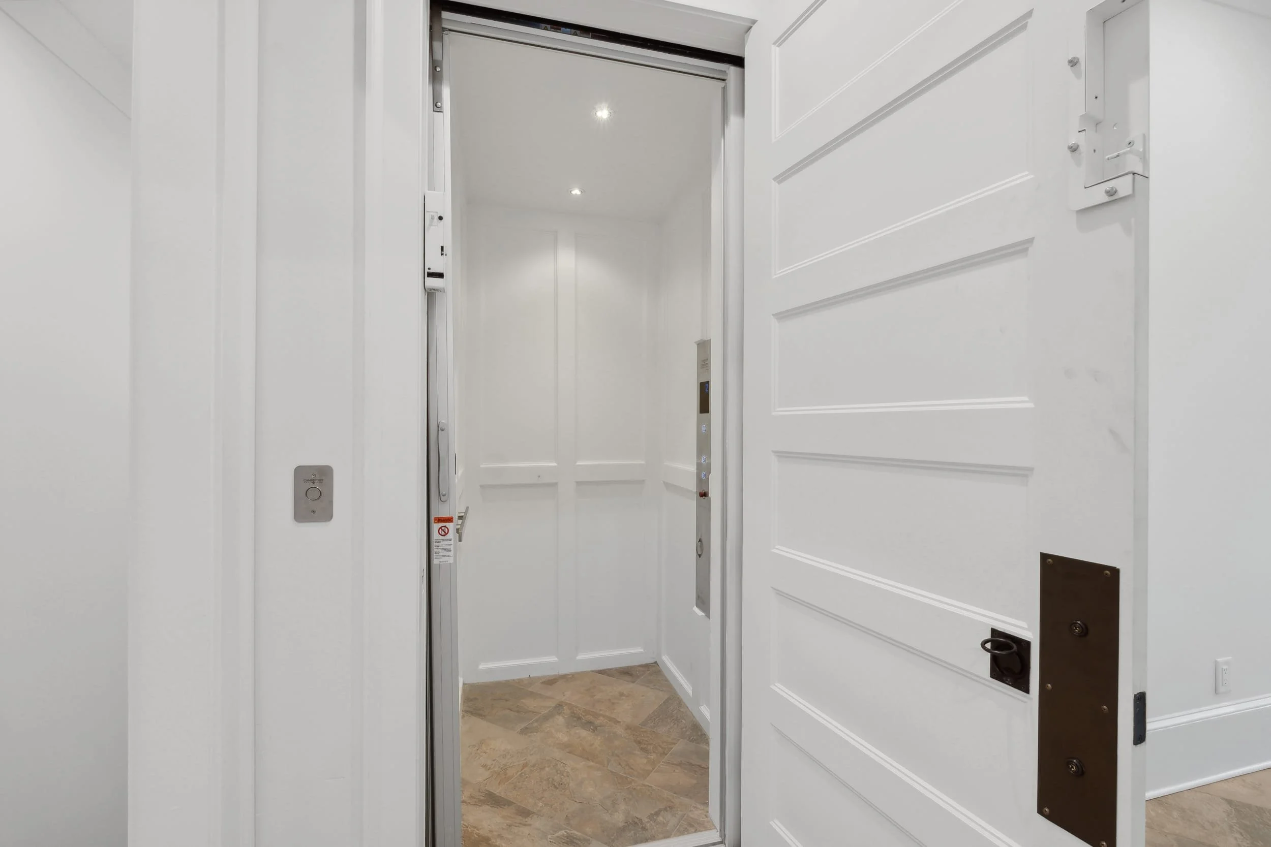 Open elevator door revealing empty elevator interior with white walls and ceiling, and a tiled floor.