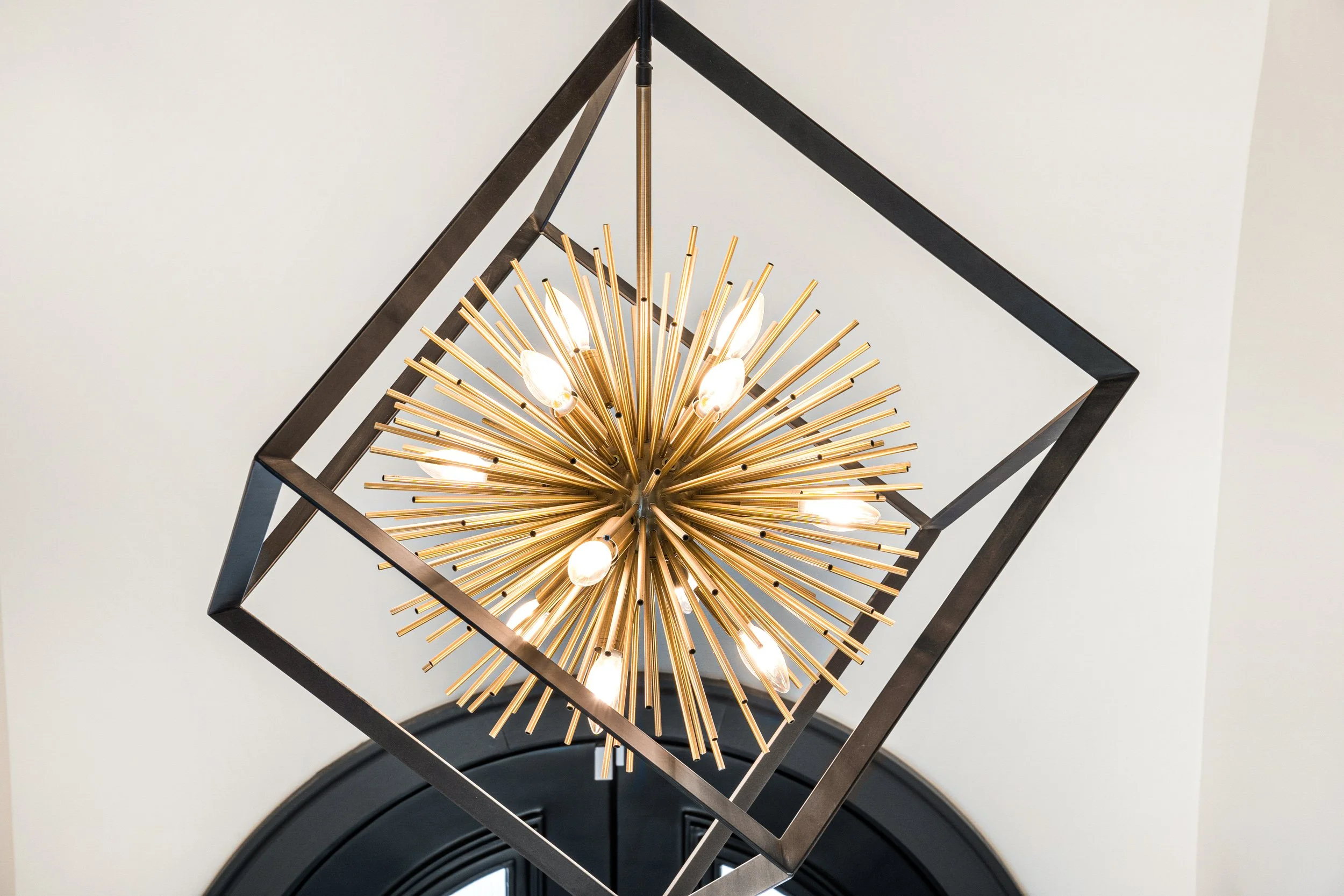 A modern chandelier with many gold rods radiating outward, framed within a black square metal structure, hanging from a white ceiling.