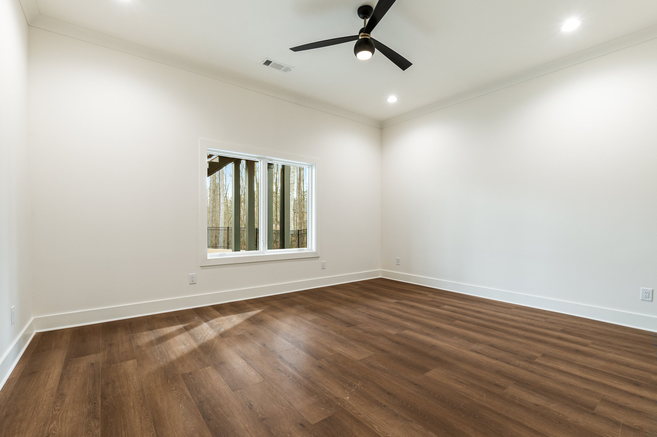Empty room with white walls, a window showing trees outside, a dark brown wooden floor, a black ceiling fan, and recessed ceiling lights.