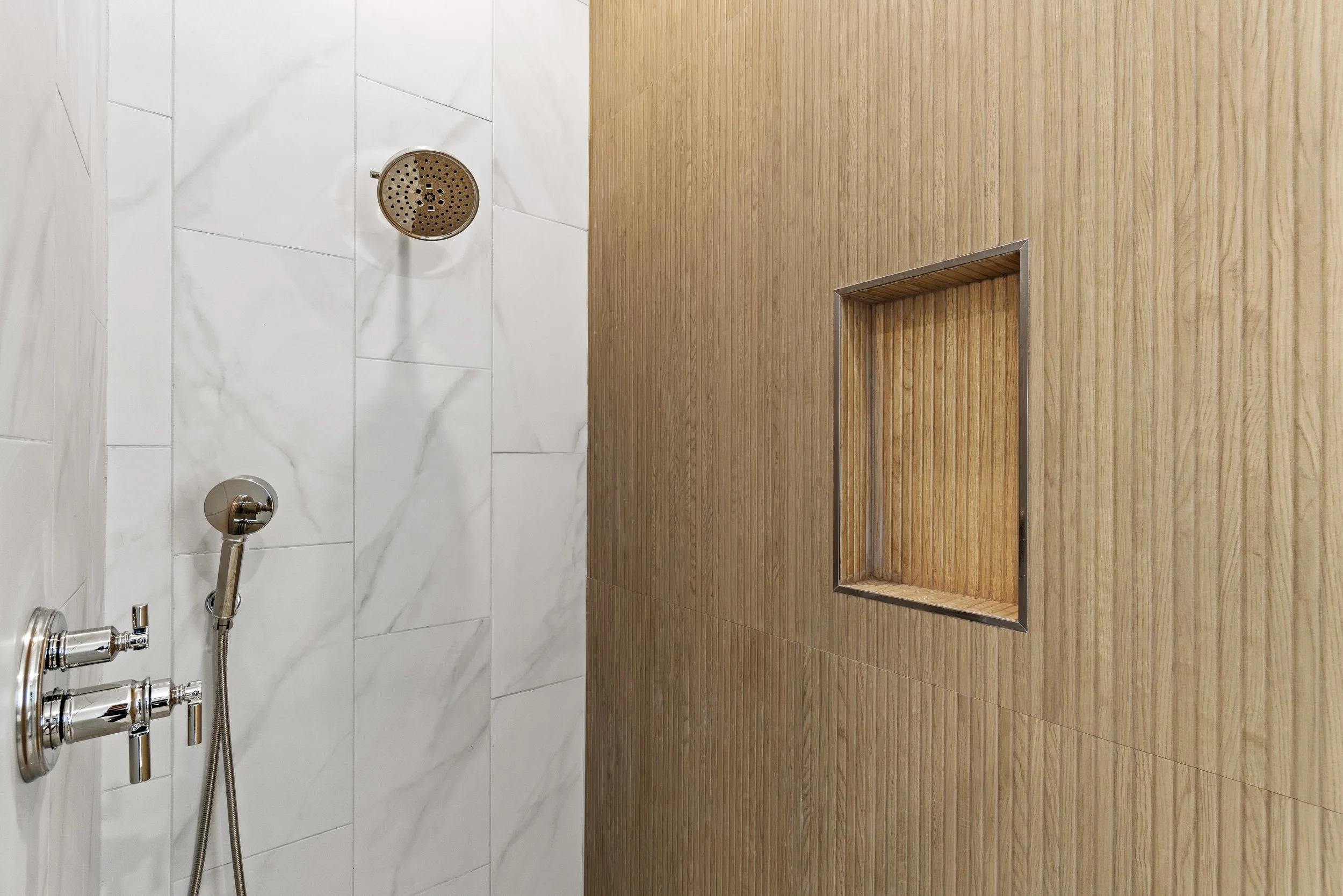 An indoor shower area with white marble tiles on the left and a wooden wall with a built-in bench on the right, featuring a ceiling-mounted showerhead and a handheld shower attachment.