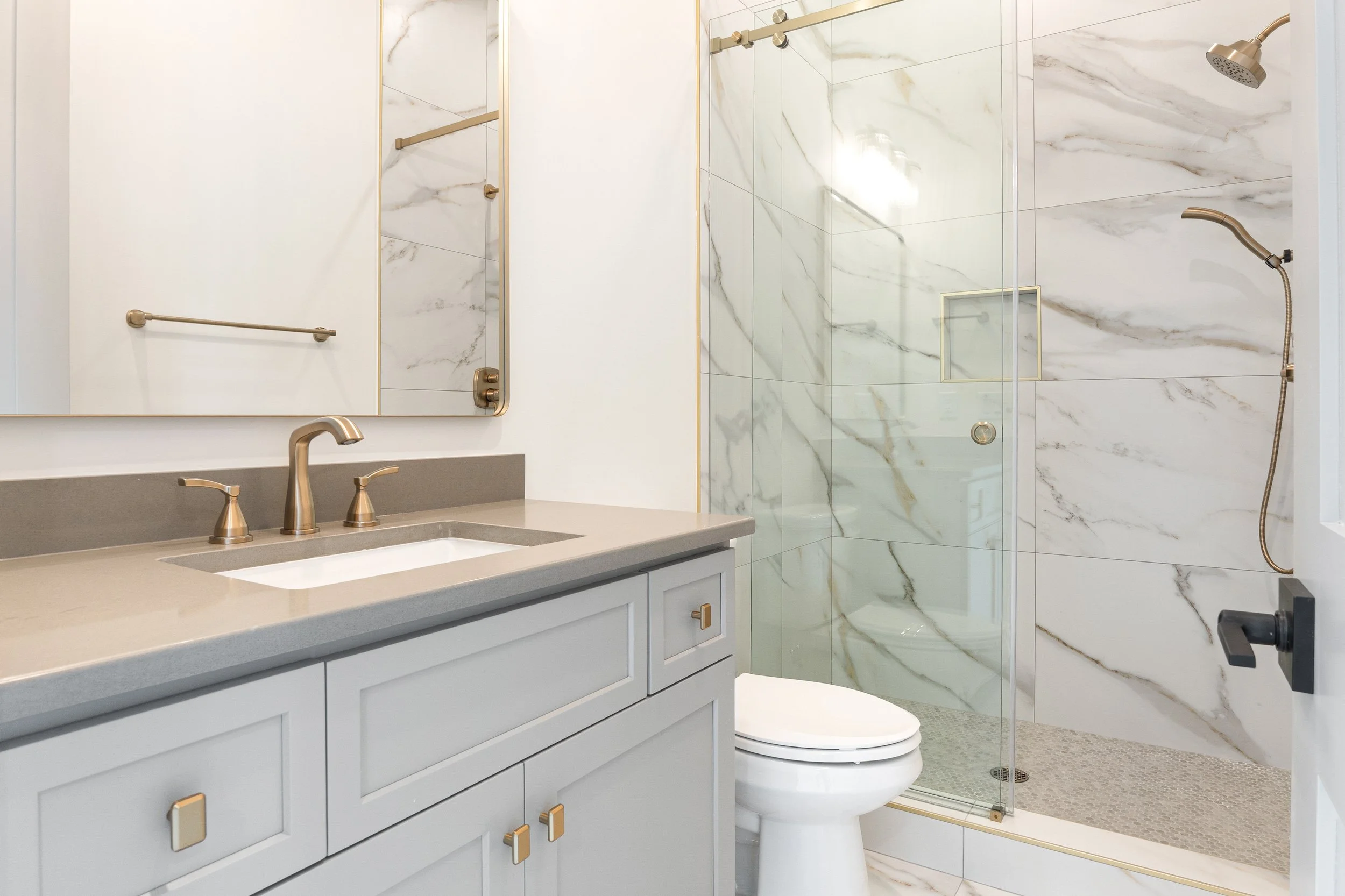 Modern bathroom with a marble shower, a white vanity with gray countertop, gold fixtures, and a mirror.