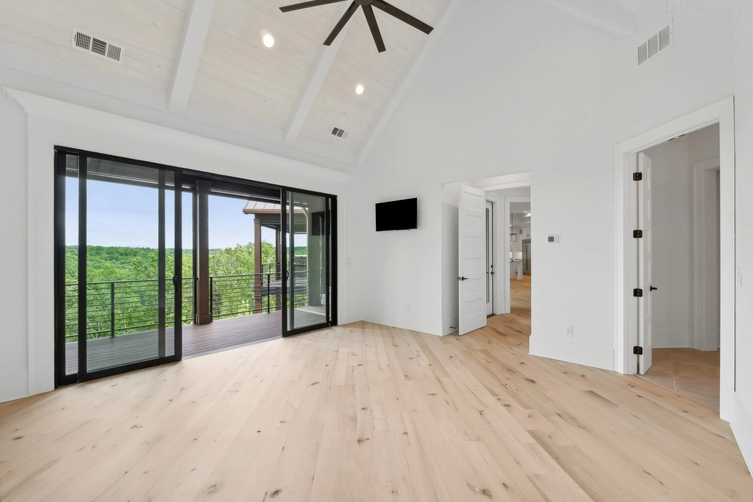 A bright living room with light wood floors, white walls, and a vaulted ceiling with black ceiling fan. Sliding glass doors lead to a balcony with lush green trees outside. There's a small black wall-mounted TV on the wall and open white doors leadin