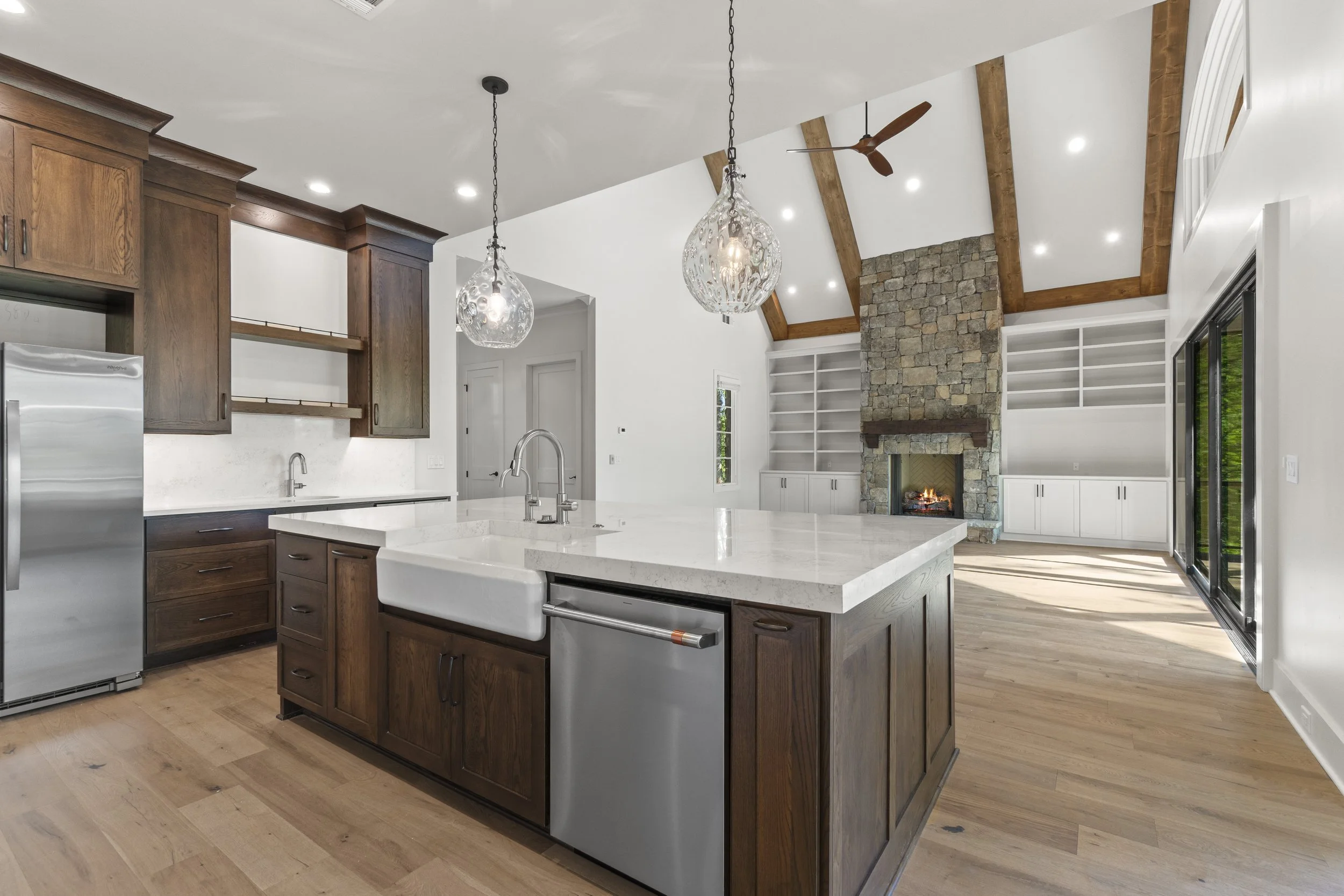 Open-concept kitchen and living room with a high vaulted ceiling, wooden beams, a stone fireplace, large windows, and modern fixtures.