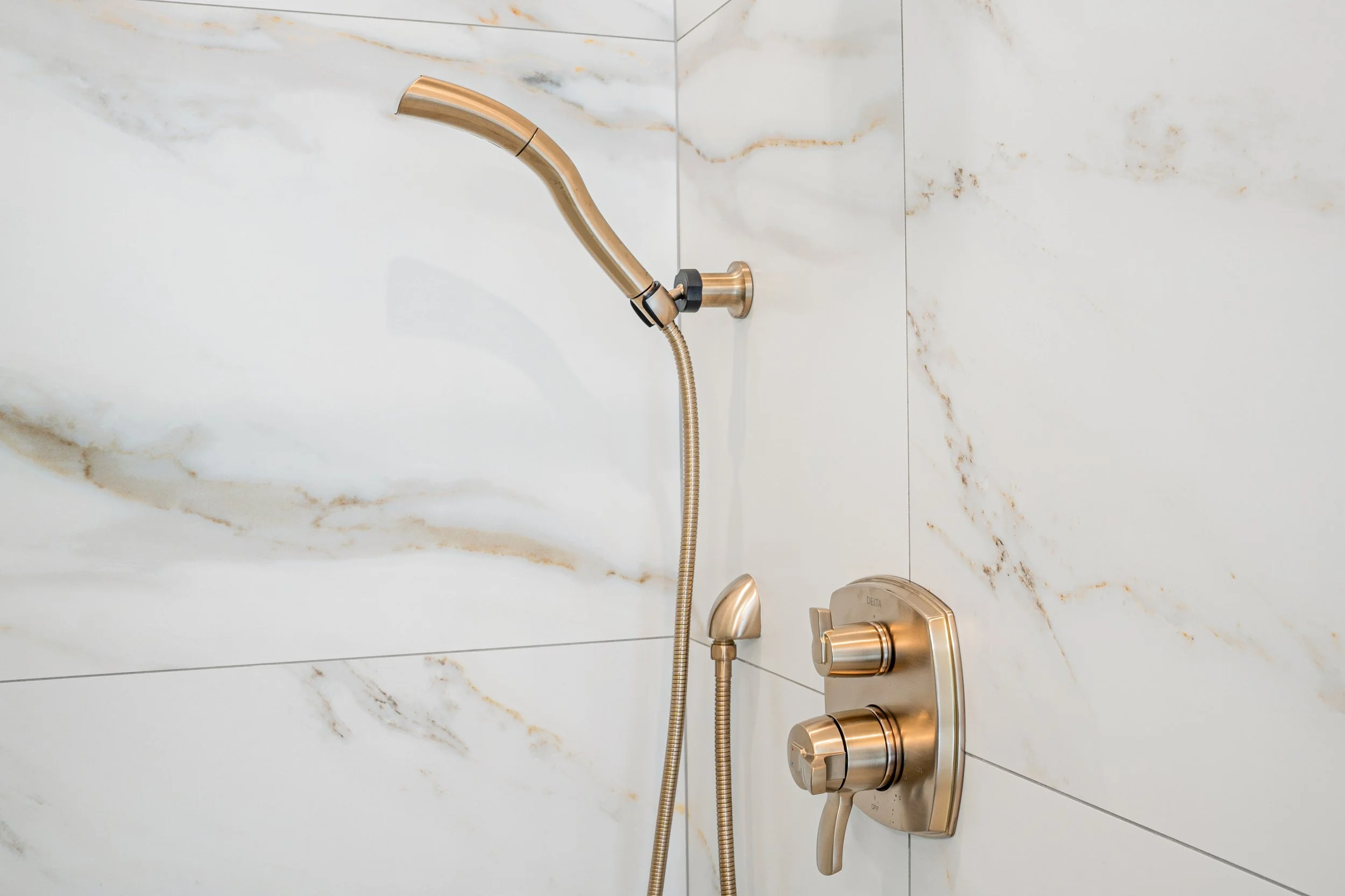 A modern shower with a gold-colored handheld showerhead mounted on a marble wall, with matching control knobs.