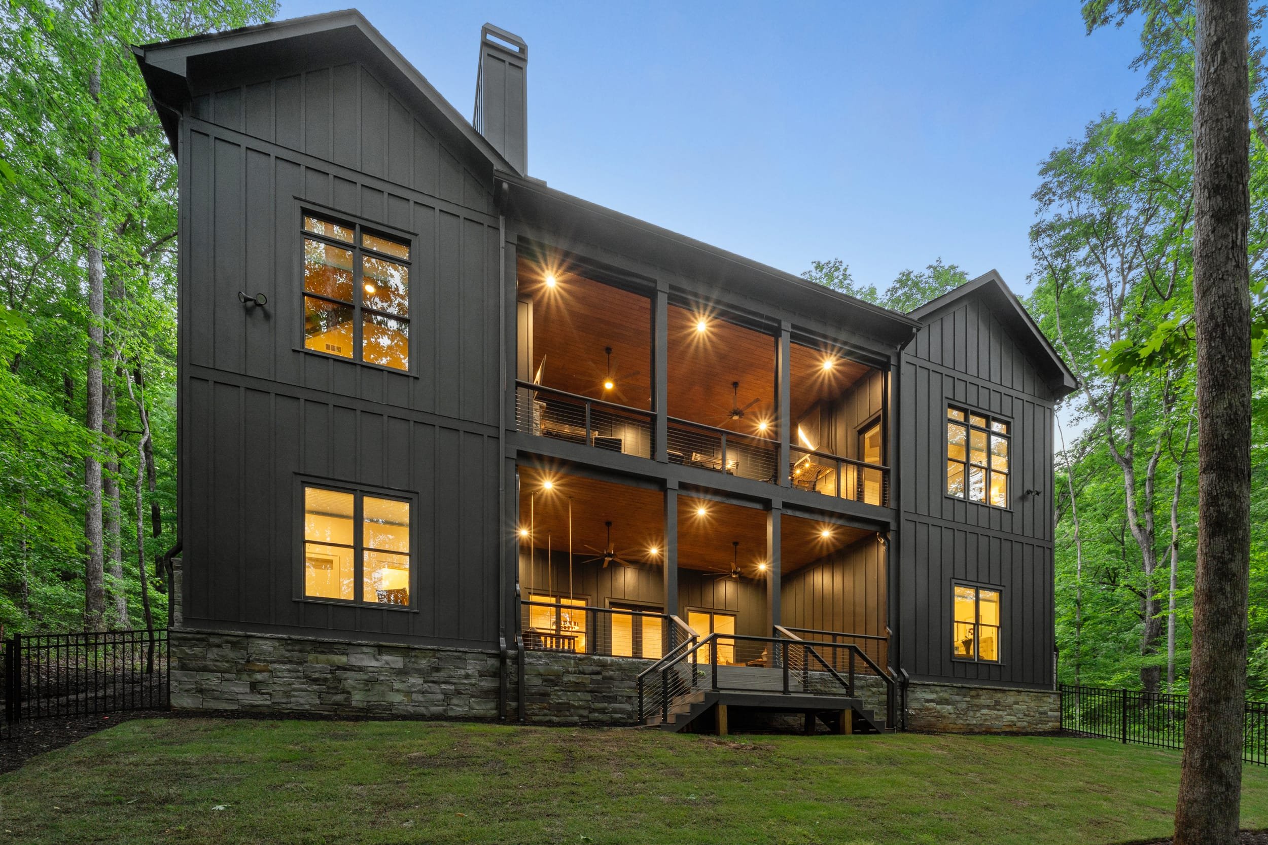 A modern multi-story house with large glass windows, illuminated from inside, surrounded by trees in a forest setting during dusk.