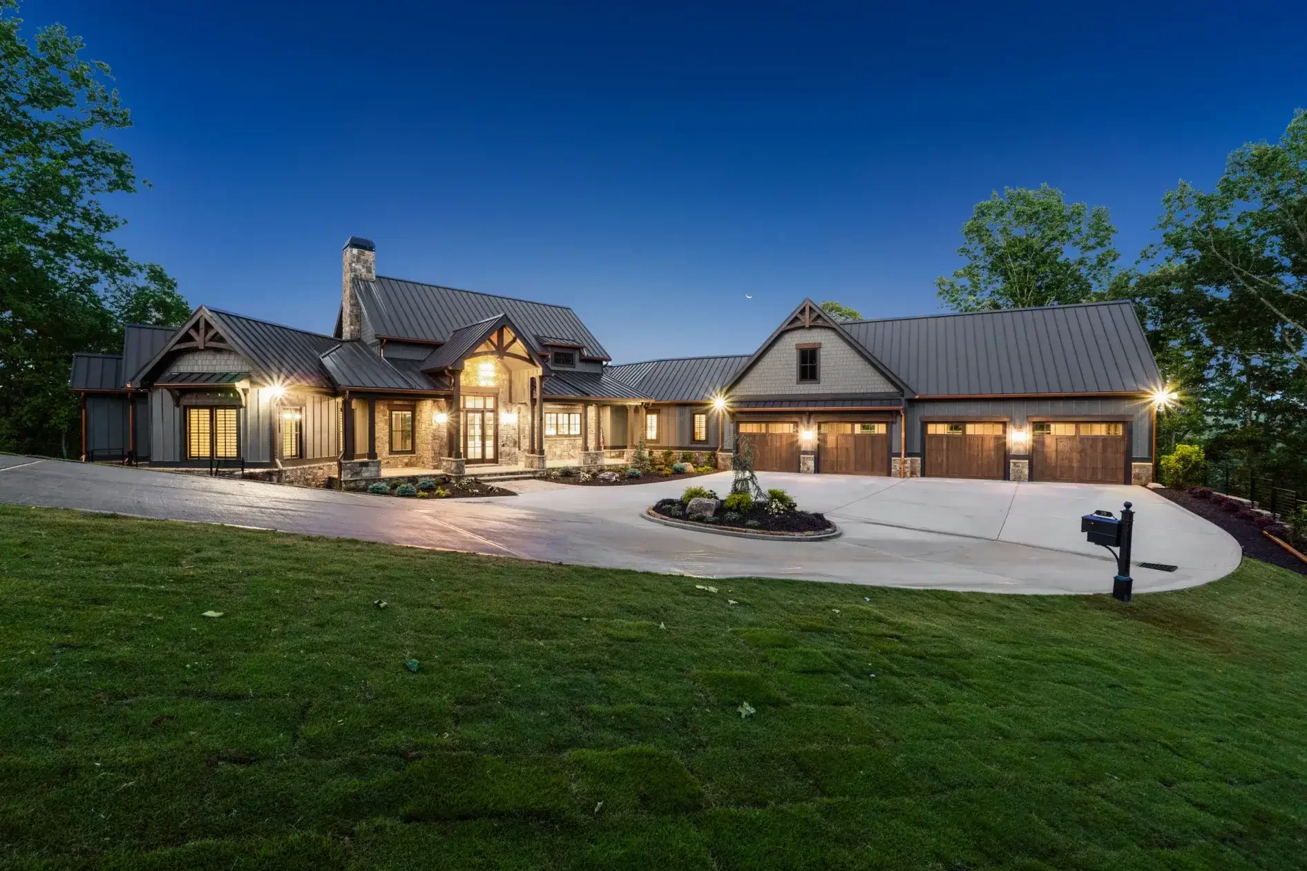 Large modern house with stone and wood exterior, metal roof, illuminated windows, surrounded by trees, with a concrete driveway, mailbox, and landscaping night scene.