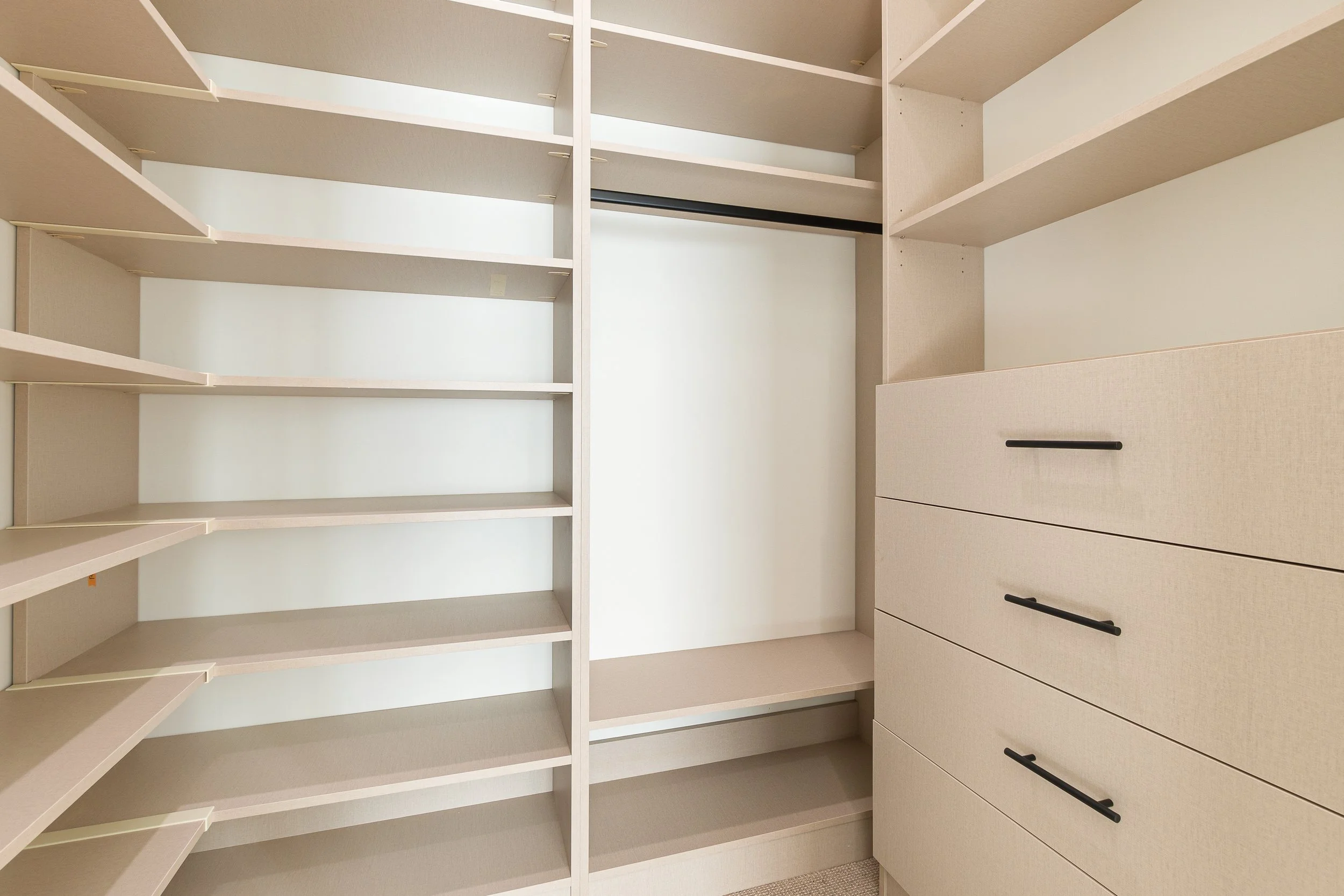 Empty walk-in closet with beige shelving and drawers, black handles, and a neutral color scheme.