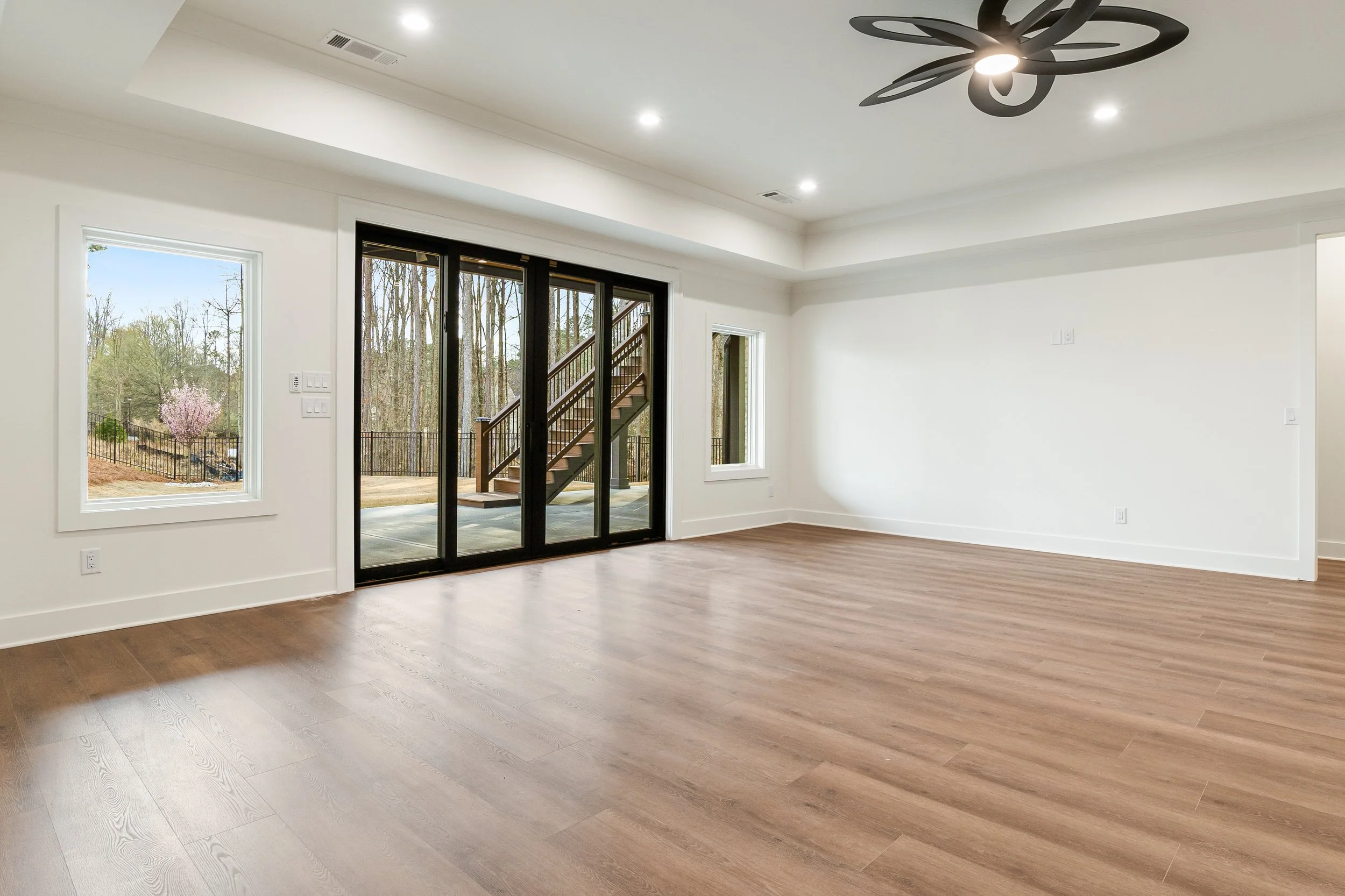 Empty living room with hardwood flooring, white walls, windows, and glass sliding door leading outside with trees and stairs visible.