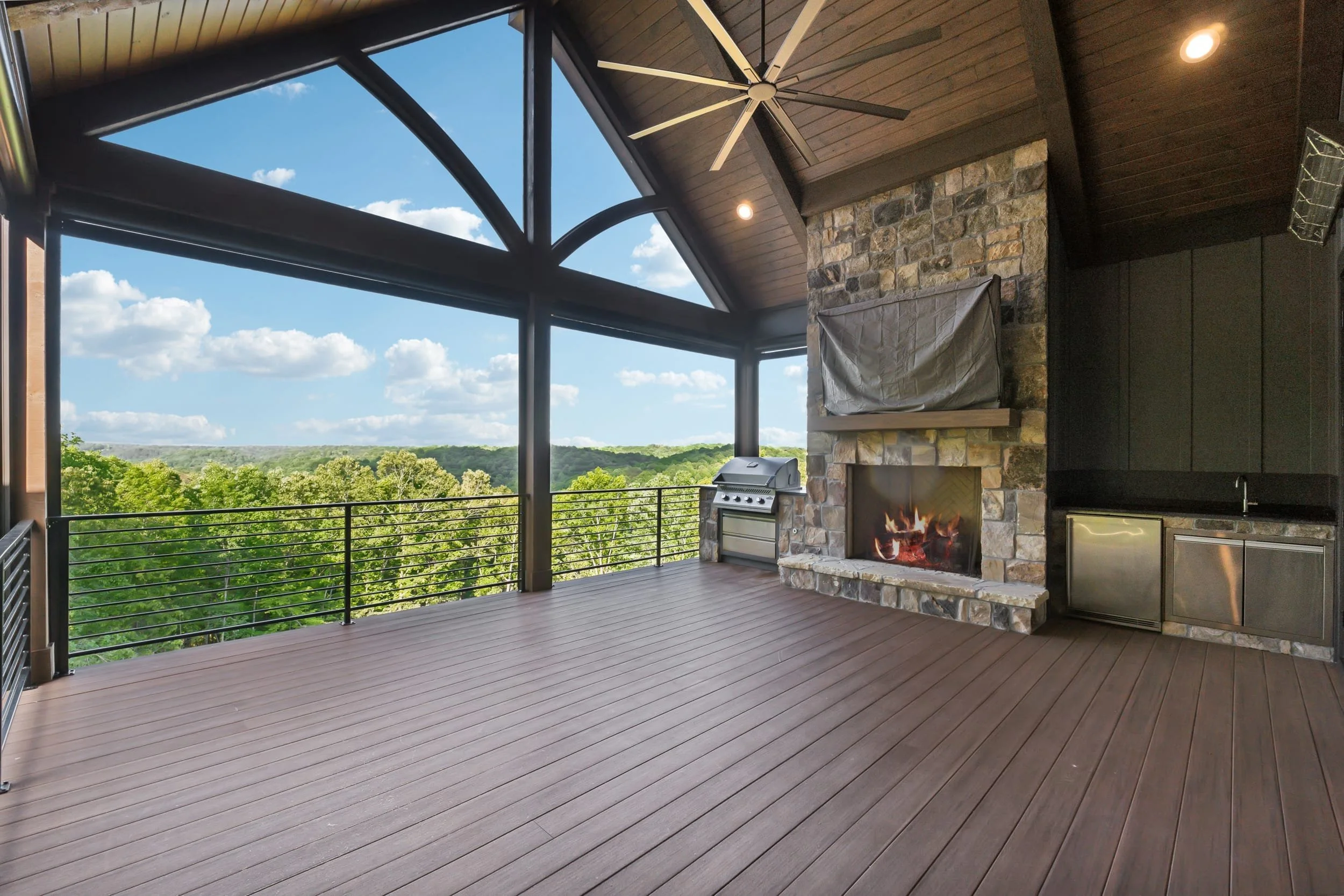 An outdoor balcony with a wooden floor and railing, a stone fireplace with a covered fireplace insert, a stainless steel grill, and a ceiling fan, with a scenic view of trees and blue sky with clouds.