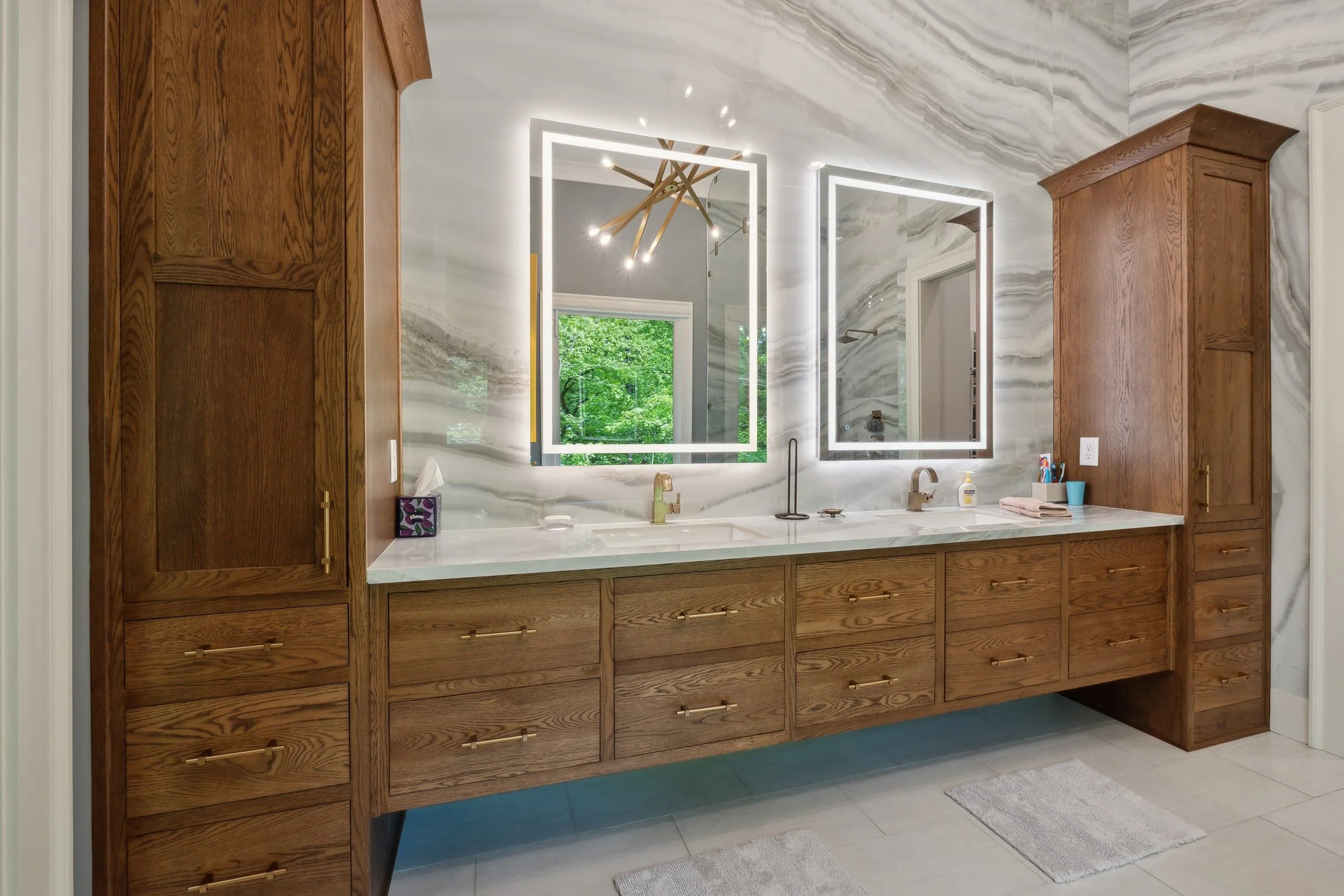 Modern bathroom vanity with wooden cabinets, a marble countertop, and double illuminated mirrors. A window with greenery outside is visible, along with some toiletries and towels on the counter.