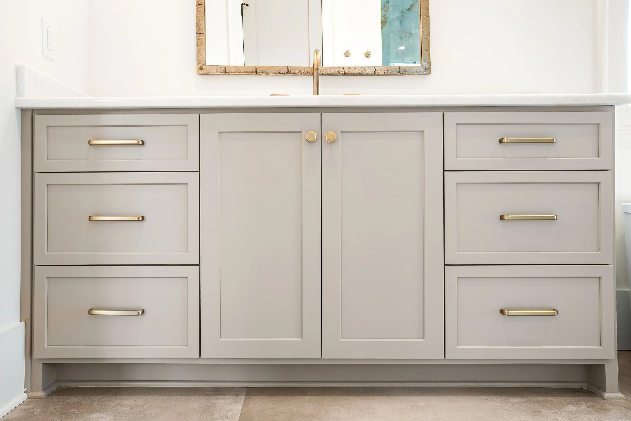 Bathroom vanity with a white countertop, white cabinets with gold handles, a large mirror with a stone frame, and a modern gold faucet.