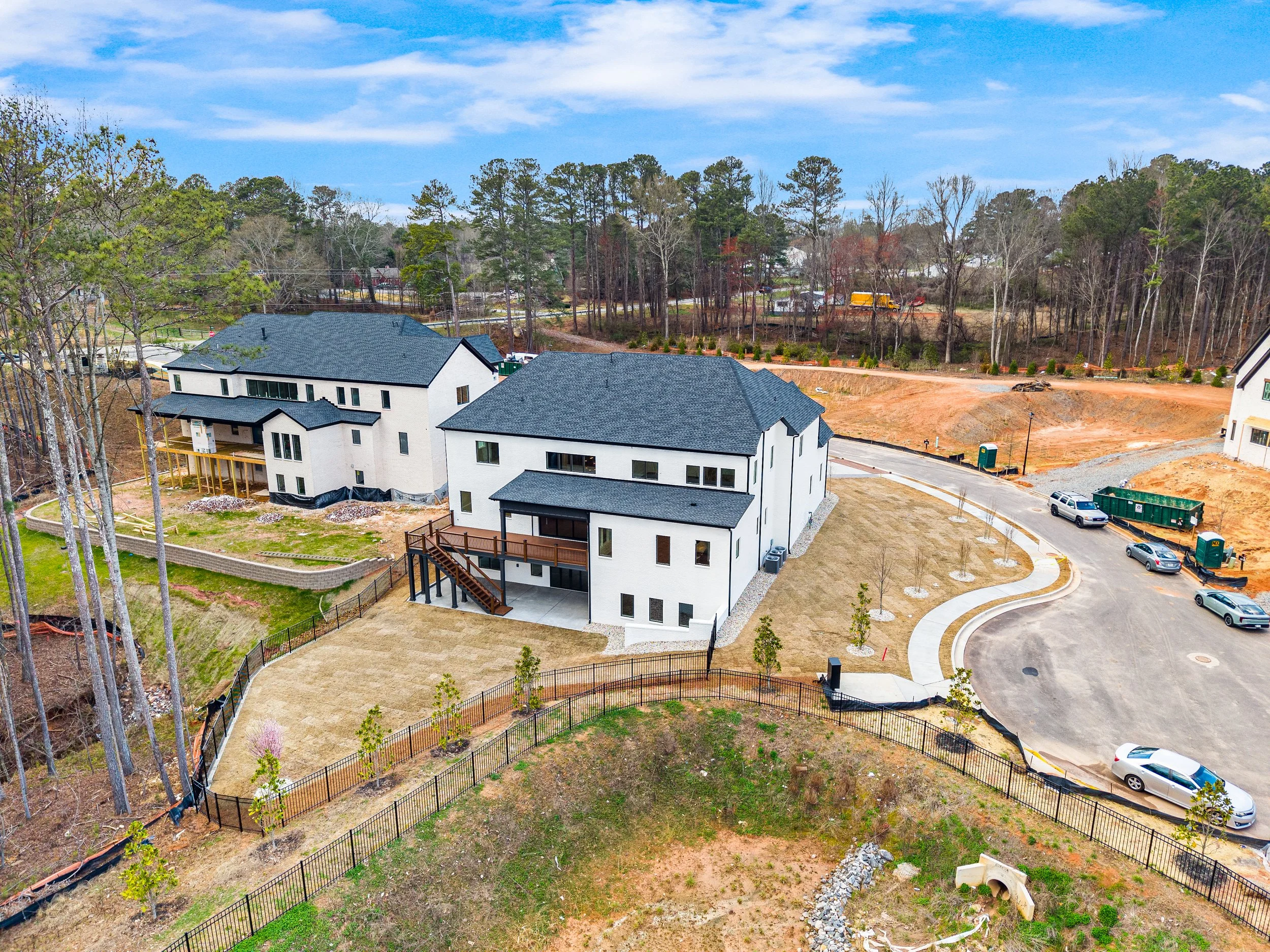 A residential construction site with two completed white houses with black roofs in the foreground, and a background of trees, dirt roads, and new development areas, under a partly cloudy sky.