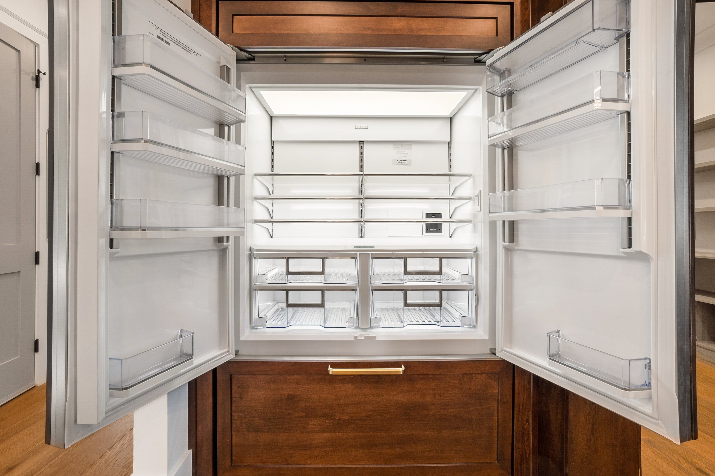 Open empty refrigerator with glass shelves and door storage compartments in a kitchen with wooden cabinets.