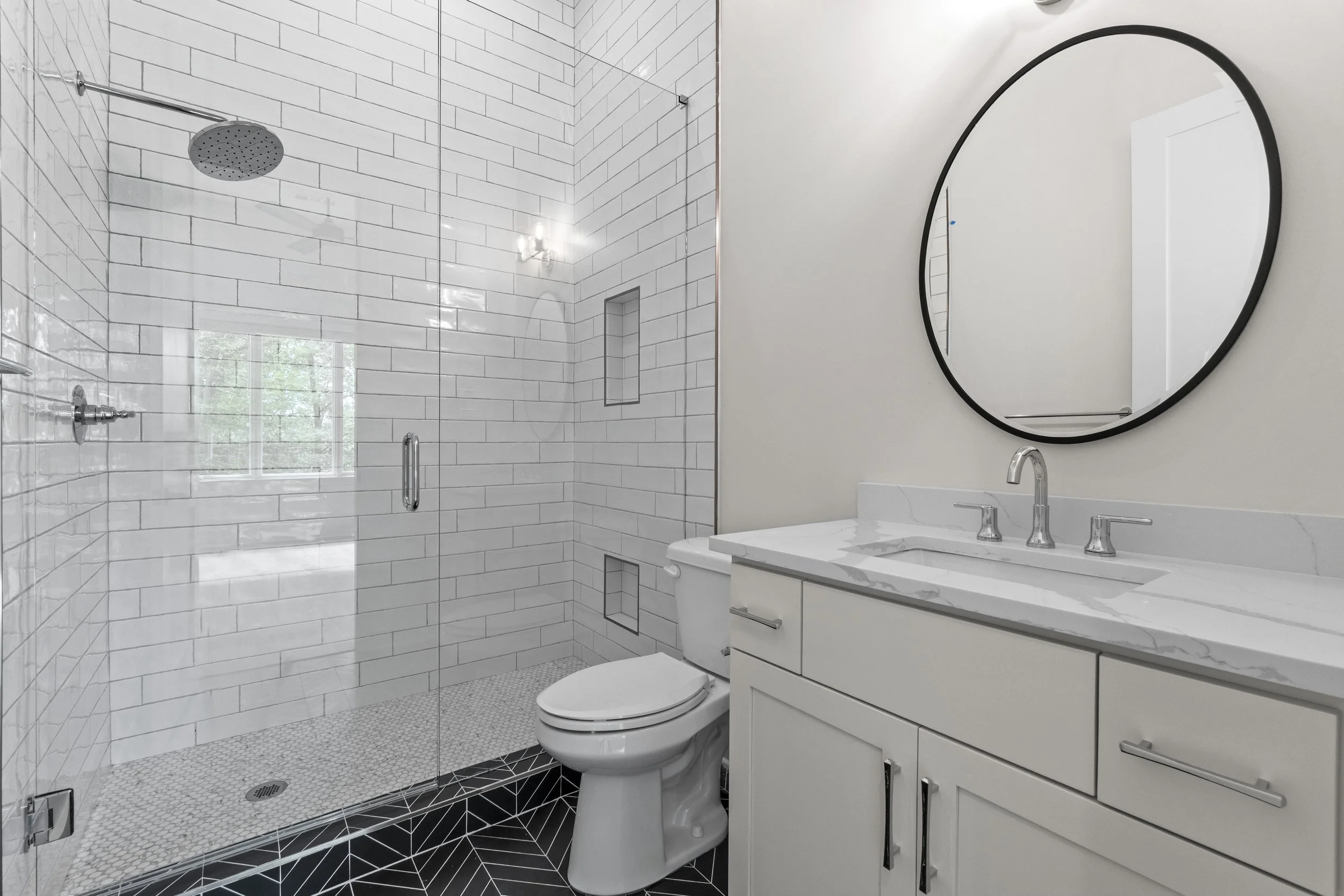 Modern bathroom with a glass-enclosed shower featuring white subway tile walls, a shower niche, and a black patterned tile floor. There is a white vanity with a marble countertop, a round mirror with a black frame, and a white toilet. A window allows