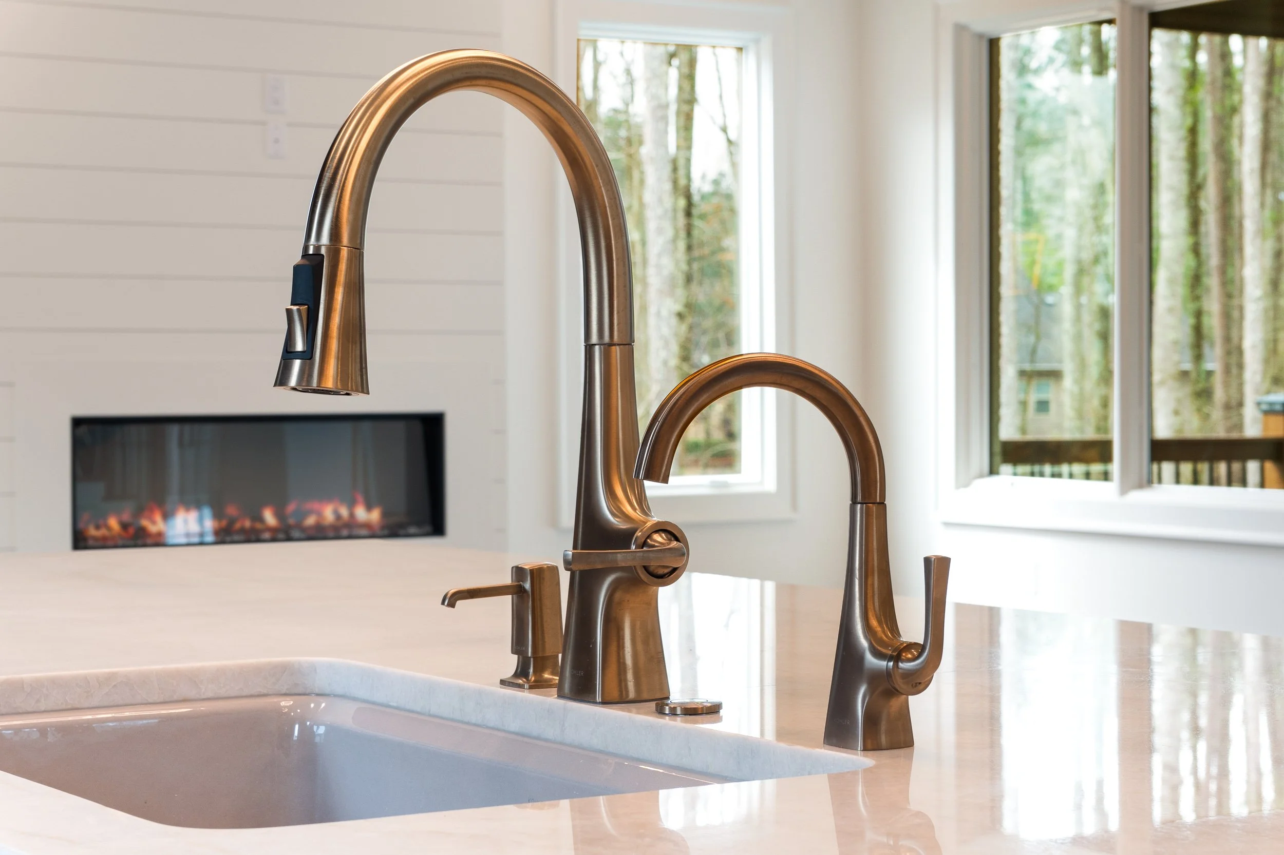 Close-up of a modern kitchen faucet on a white countertop with a sink, with large windows showing trees outside and a fireplace in the background.