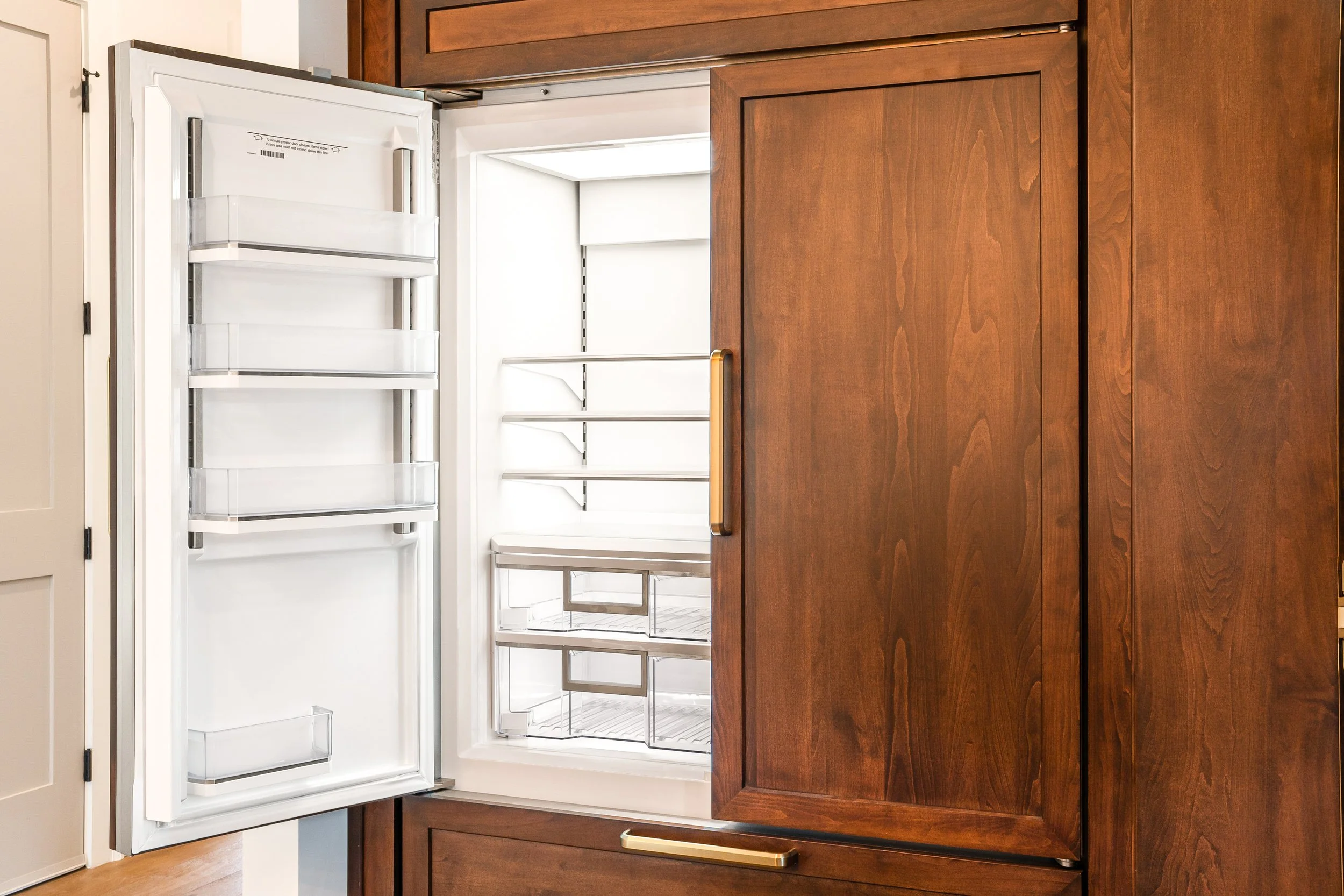 Empty open refrigerator with white interior, glass shelves, and a wooden cabinetry surrounding.