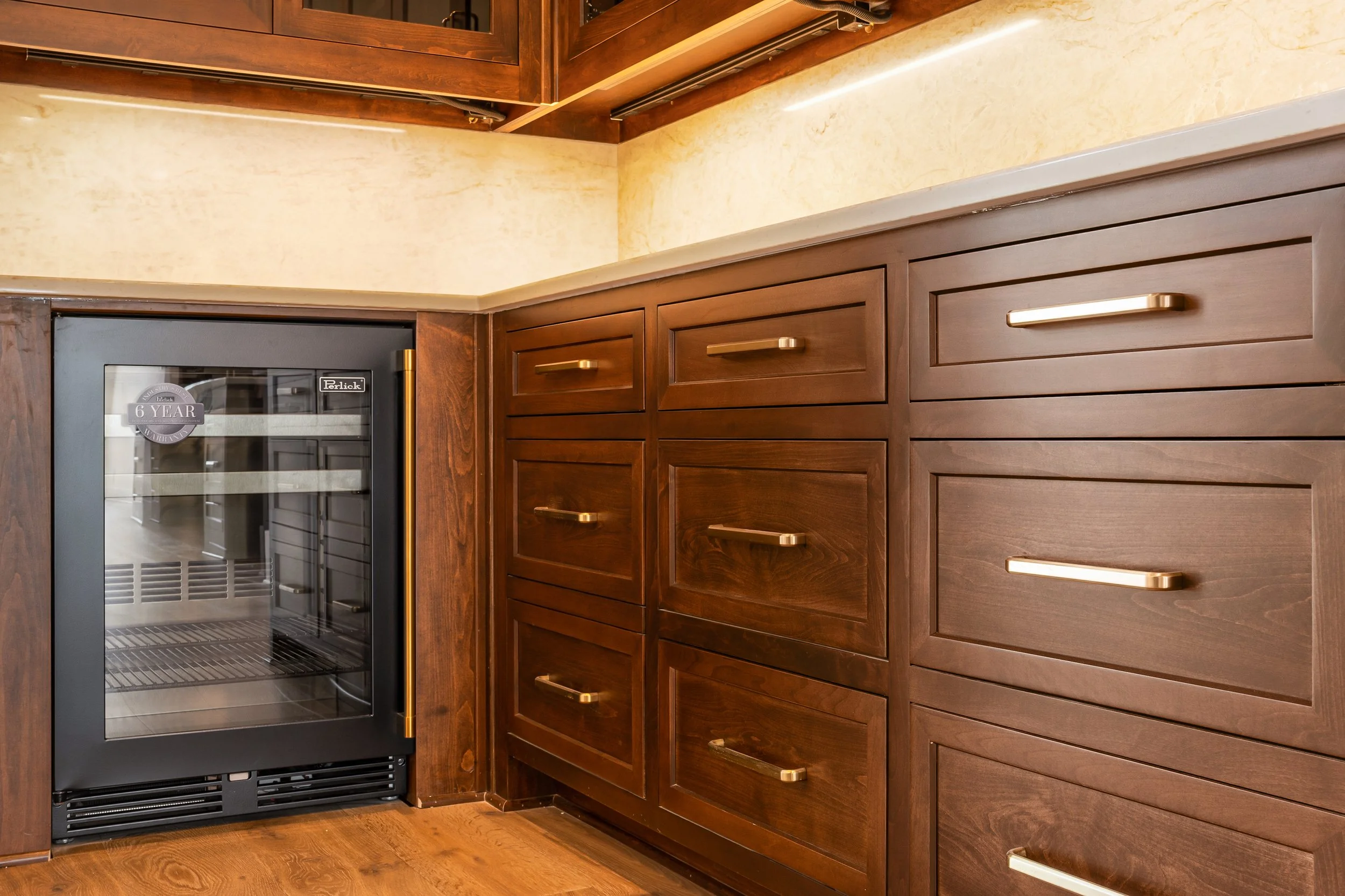A kitchen corner with wooden cabinets and a small wine refrigerator. The cabinets are dark brown with sleek gold handles, and the wine fridge has a glass door with a 