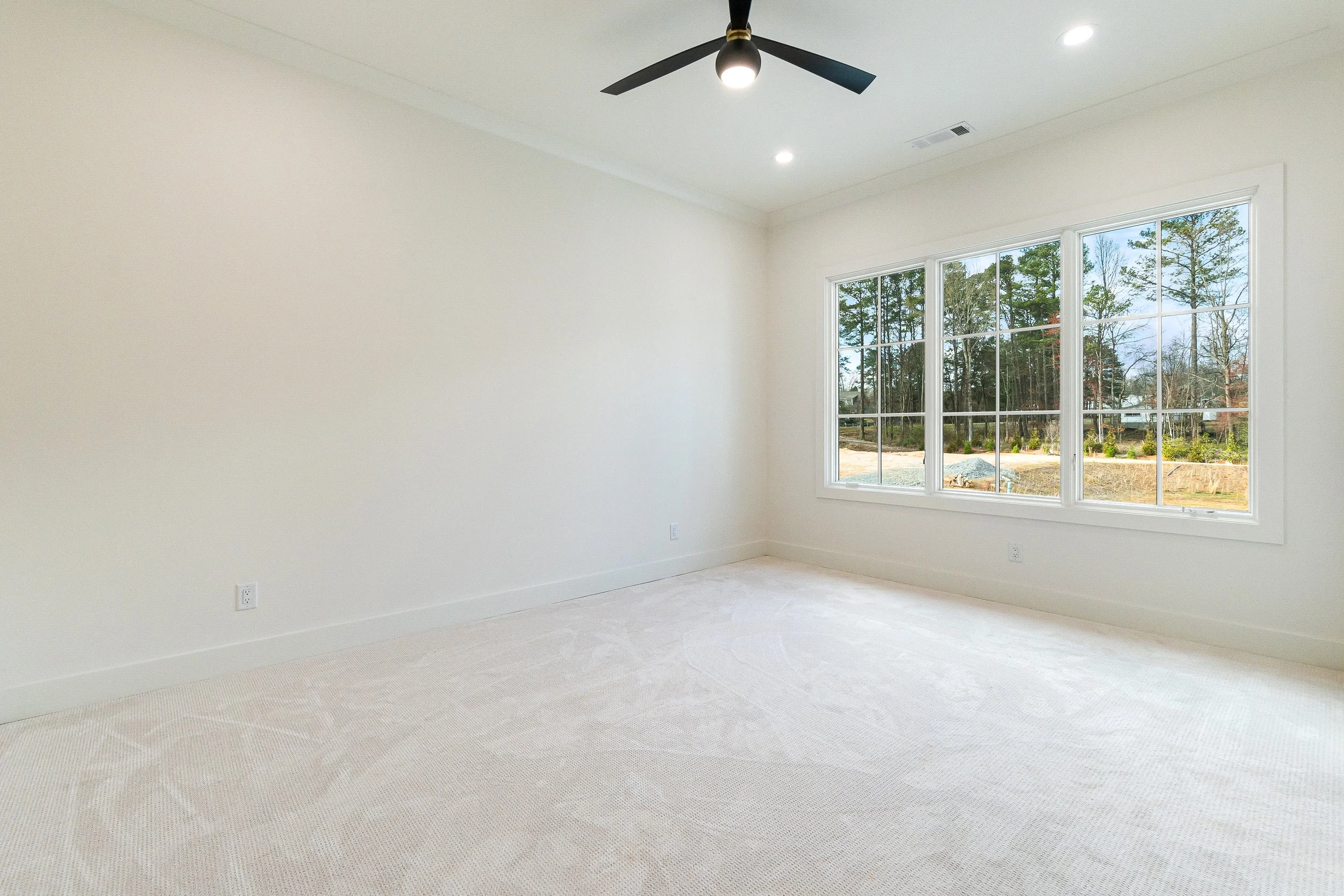 Empty room with large window showing trees and a yard outside, white walls, ceiling fan with three blades, and carpeted floor.
