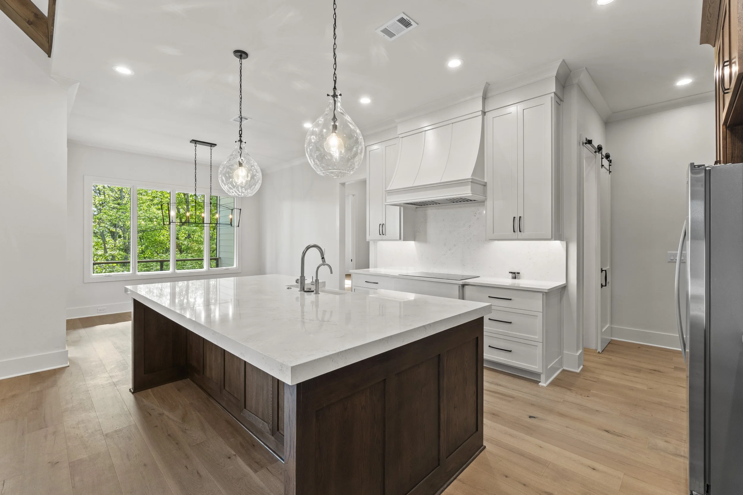 Modern kitchen with white cabinets, a white marble island, pendant lights, and a large window showing greenery outside.