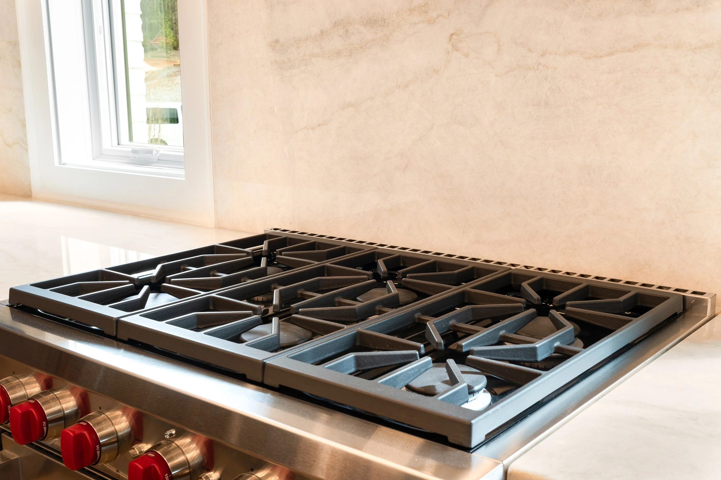 Close-up view of a stainless steel gas stove with multiple burners and red control knobs, situated in a modern kitchen with a beige countertop and a window in the background.
