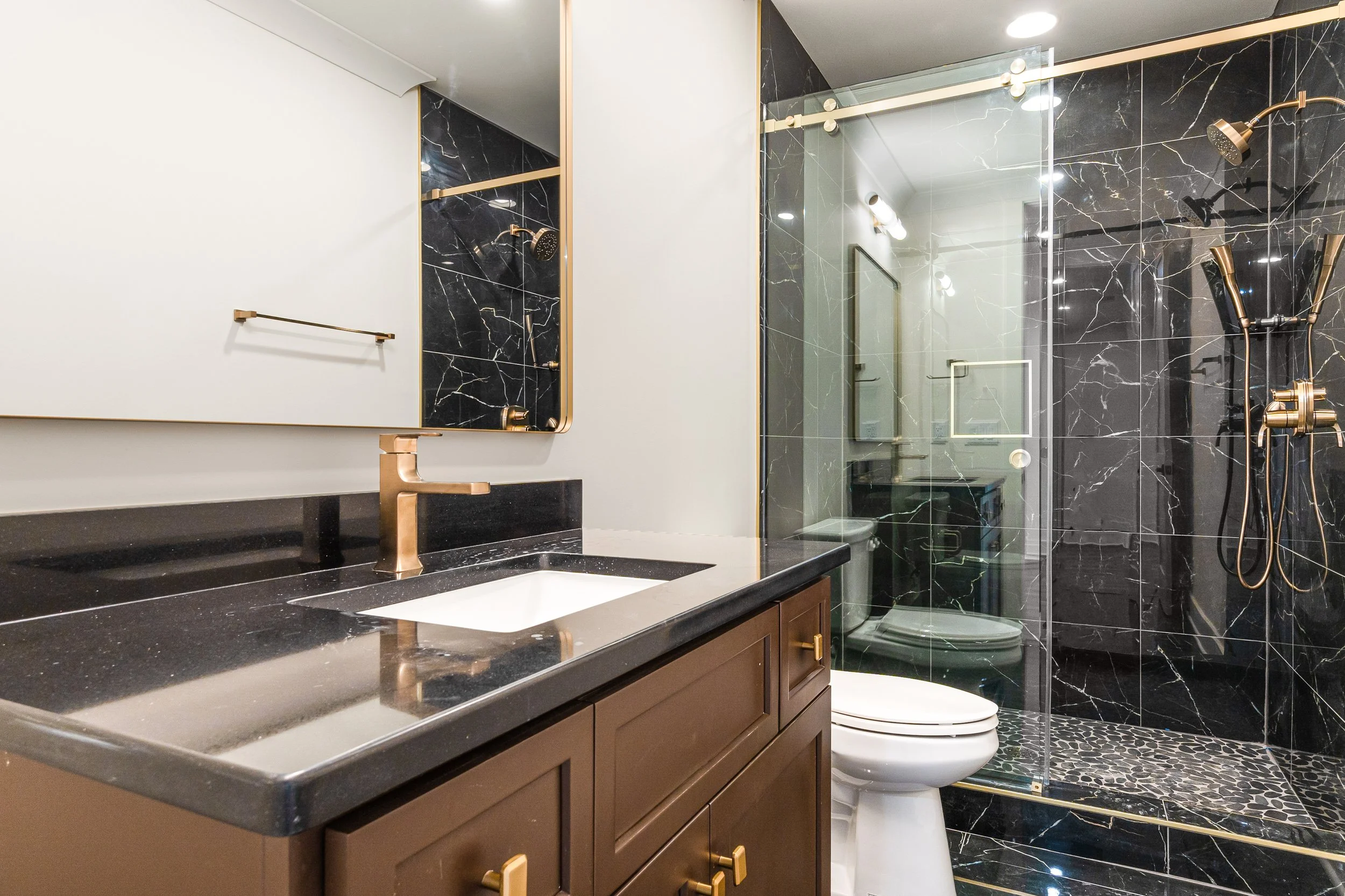 Modern bathroom with black marble tiles, a glass shower enclosure, a white toilet, a vanity with a black countertop, a gold faucet, and gold hardware, illuminated by ceiling lights.