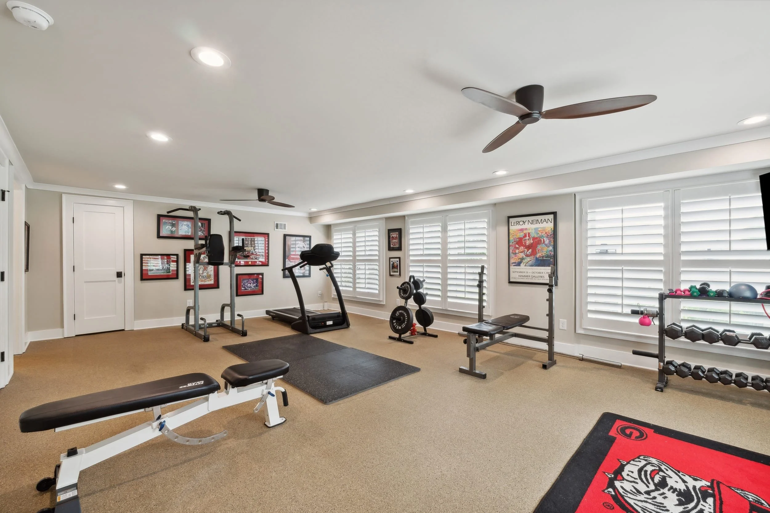 A home gym with exercise equipment including a treadmill, weight bench, dumbbells, and a pull-up bar in a room with large windows, framed artwork, and a ceiling fan.